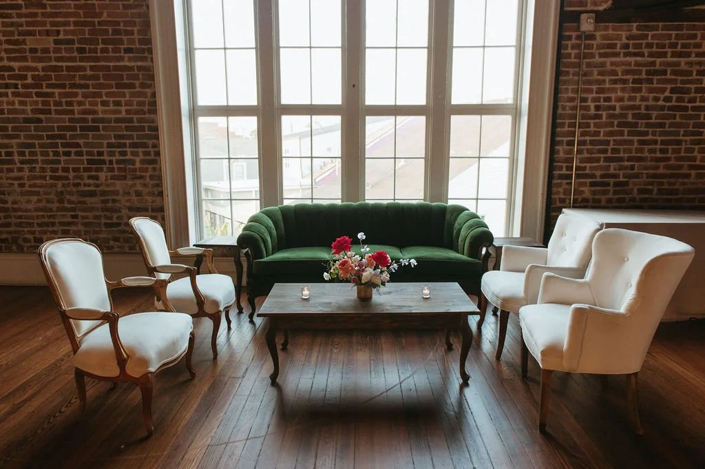 Cozy sitting area with a green velvet sofa, white armchairs, a wooden coffee table with a flower vase, and candles, in front of a large window with brick walls.