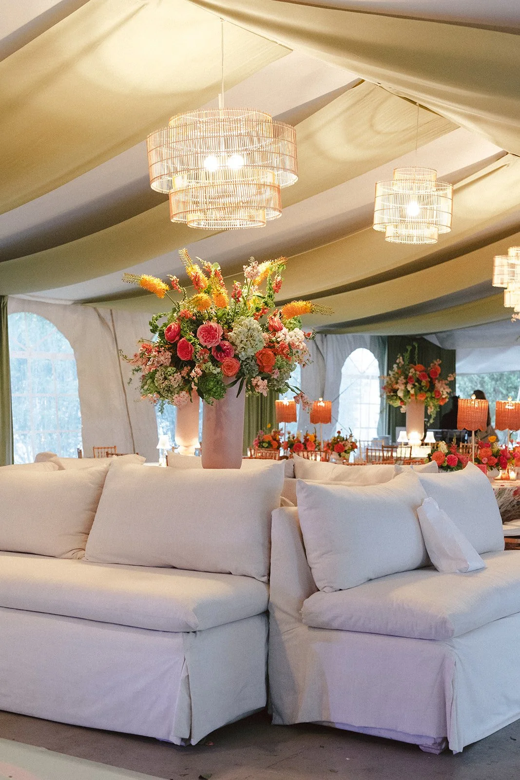 Elegant event space decorated with large floral arrangements in tall vases, cream-colored sofa, and soft lighting from chandelier and lamps.