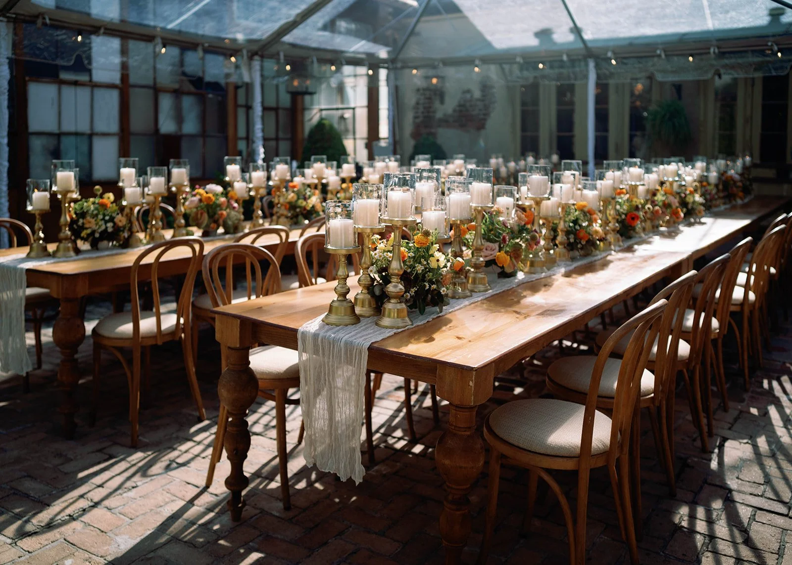 Decorated long wooden dining table with white pillar candles in glass hurricanes, floral arrangements with orange, yellow, pink, and white flowers, set in a glass-enclosed outdoor or greenhouse space with string lights overhead.