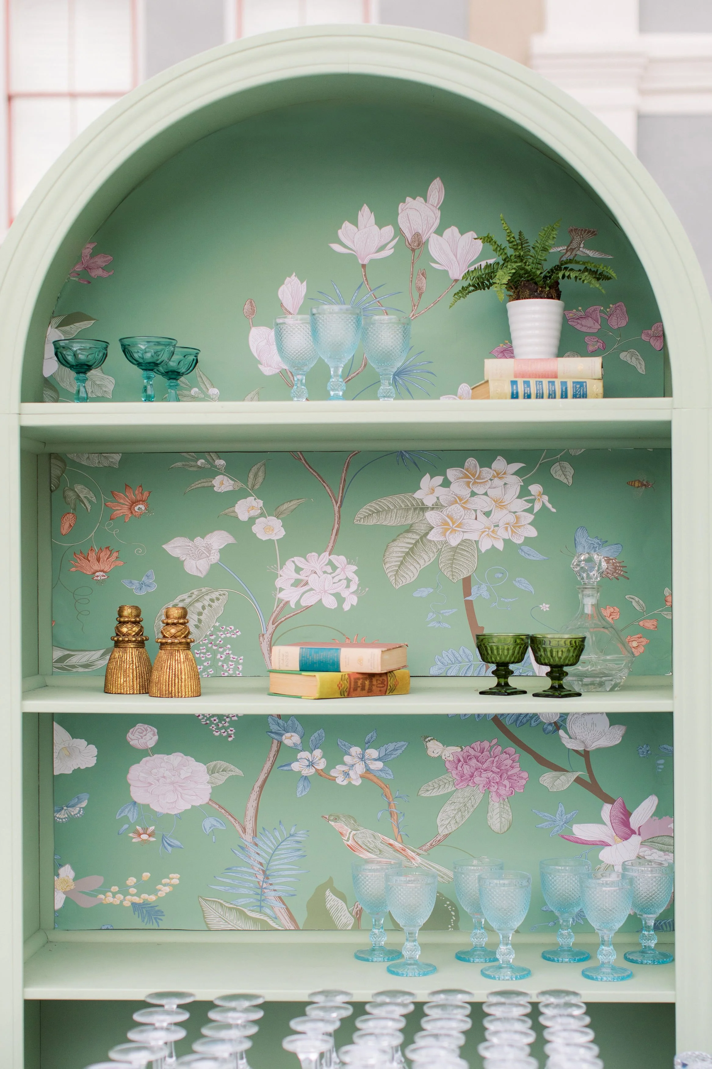 A green display shelf with four tiers, decorated with floral wallpaper featuring flowers and birds. The top tier holds four light blue glasses, some books, and a potted fern. The middle tier displays two brass bottles, several books, and some green g