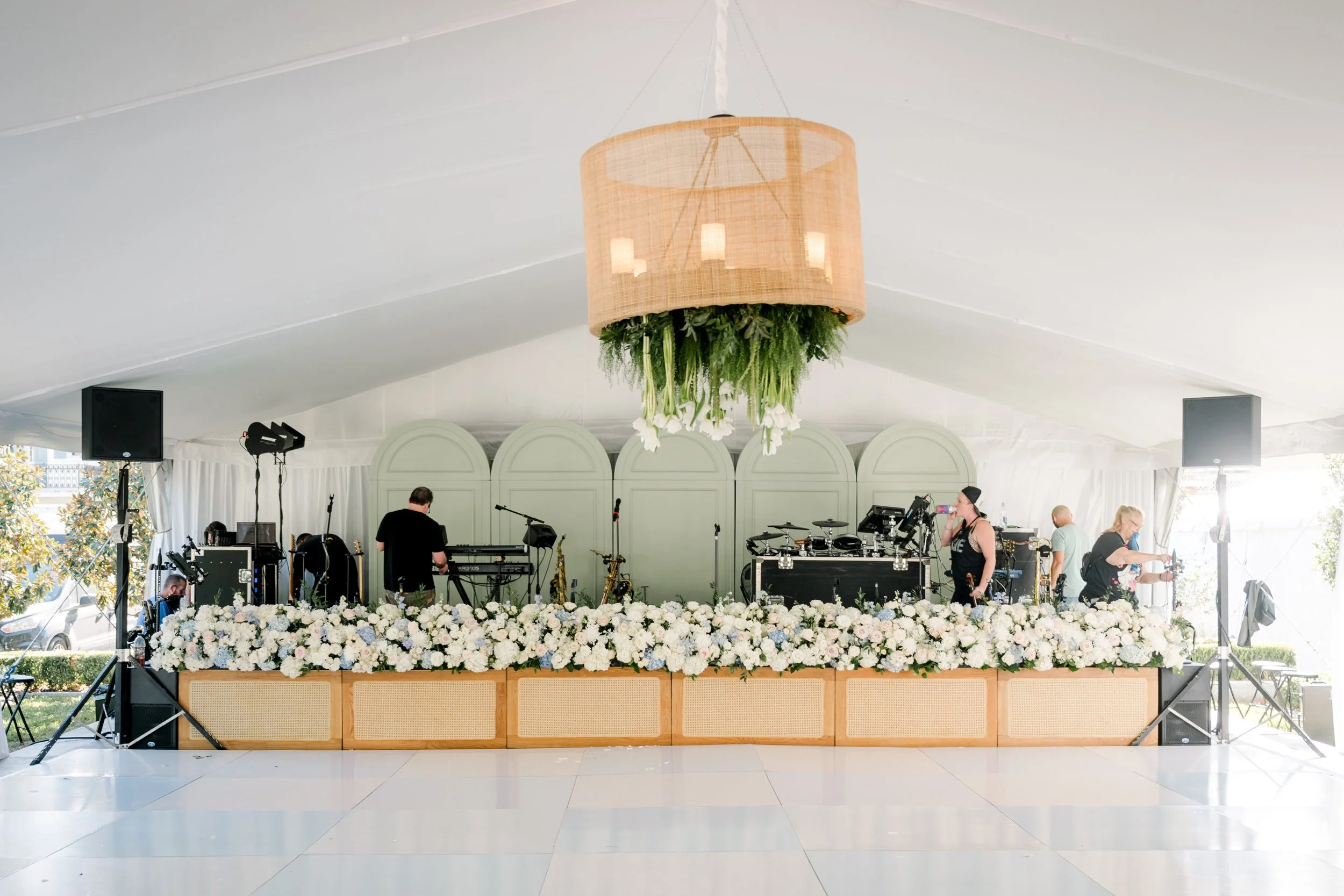 A music stage decorated with white flowers and greenery, set under a white tent with a large pendant light hanging from the ceiling. People are preparing for a performance with musical instruments and sound equipment visible.