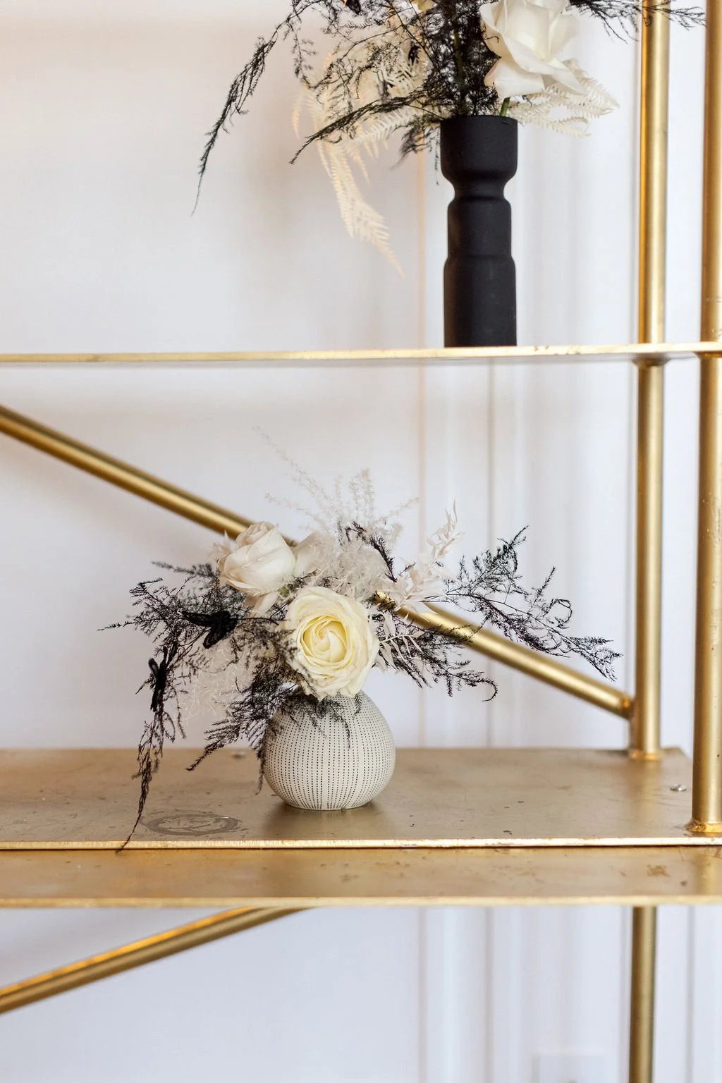 A decorative arrangement of white roses and dark foliage in a small, textured white vase, placed on a gold and glass shelf with a black vase containing white flowers and foliage behind it.