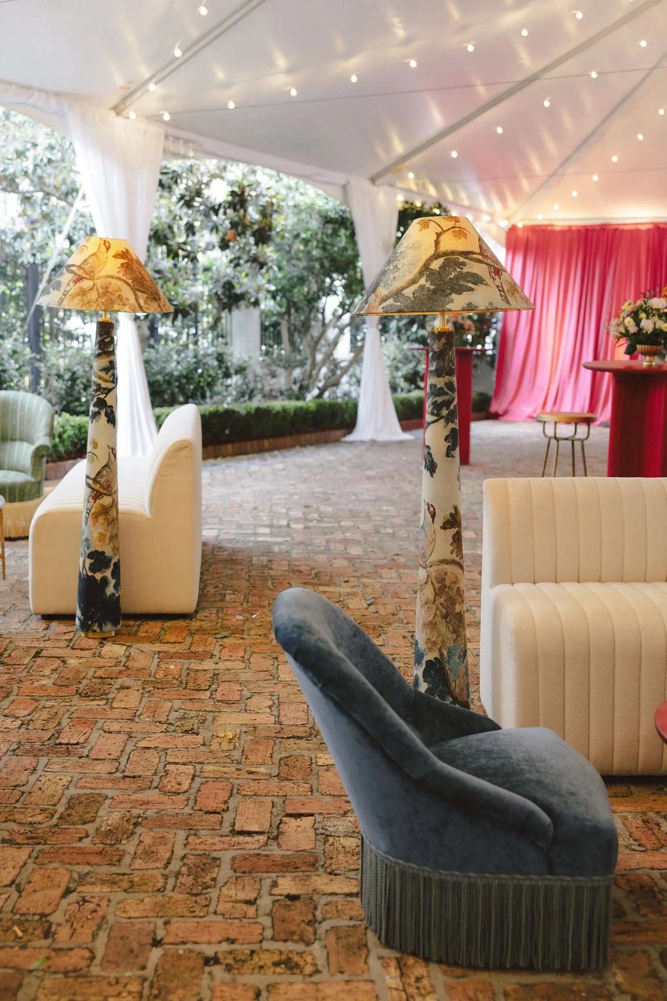 Decorative lamps, white sofas, a red bar with a bouquet of flowers, and a pink curtain inside a tented outdoor setting with string lights and trees in the background.
