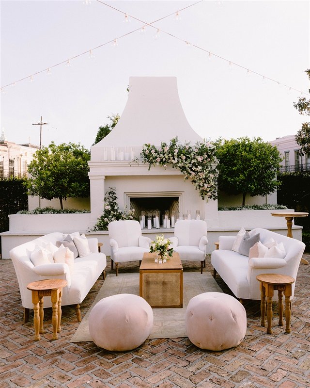 Elegant outdoor seating area with white sofas and chairs arranged around a fireplace with floral decorations, set on a brick patio with string lights overhead.