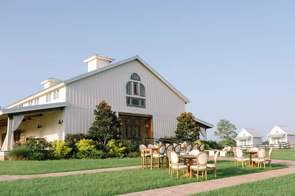 A large white barn-style building with outdoor seating and a grassy lawn, with smaller white houses in the background under a clear blue sky.