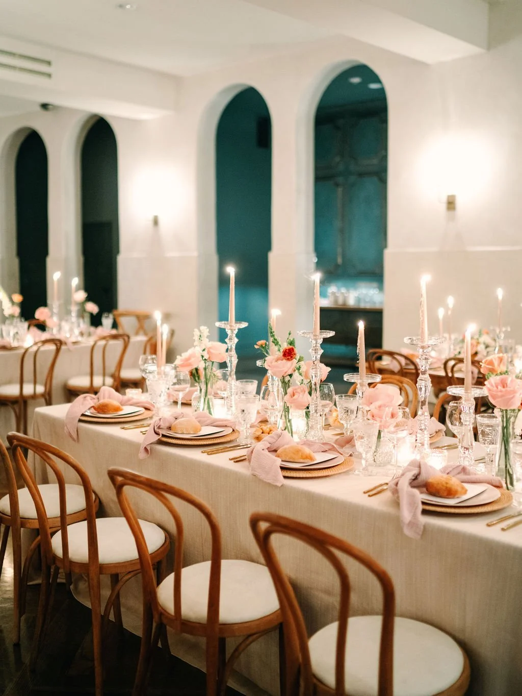 Elegant dining table setup with pink and white floral arrangements, lit candles, and place settings with bread rolls, in a decorated ballroom.