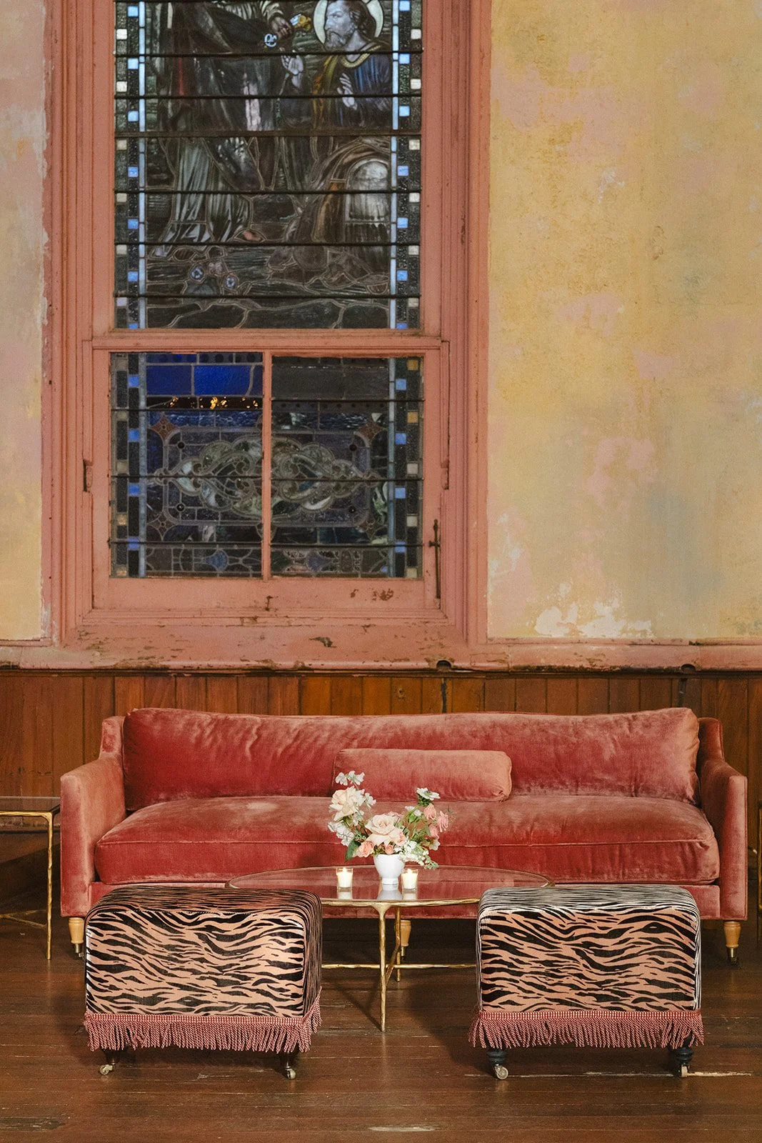 A vintage interior with a pink velvet couch, a small glass-top table with a bouquet of flowers, and two zebra-patterned ottomans, in front of a stained-glass window and a yellowed wall.