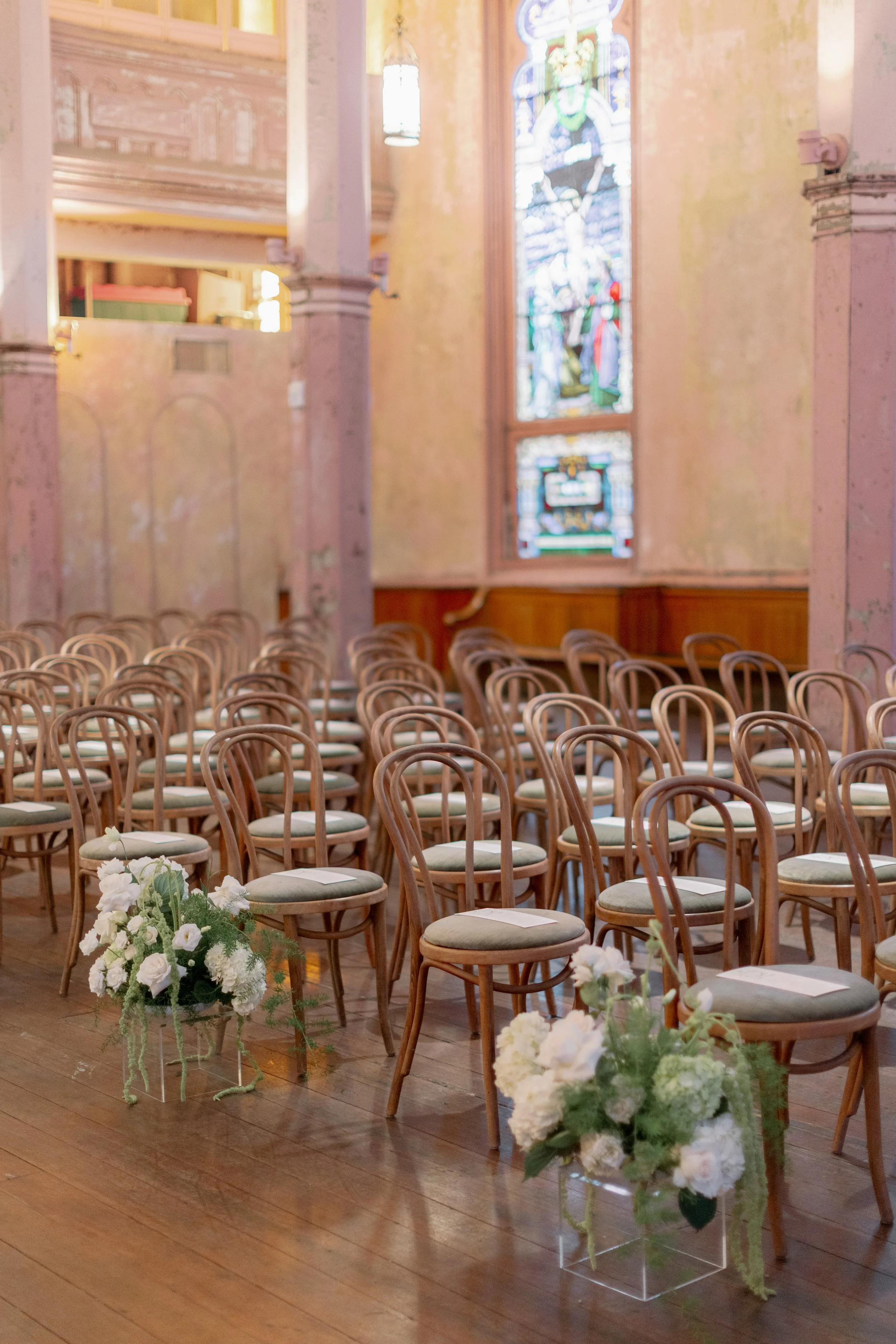 Interior of a church set up for a wedding, with rows of wooden chairs, floral arrangements in vases, stained glass windows, and soft lighting.