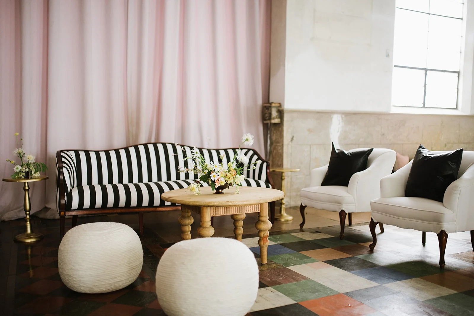 A cozy living room with vintage and modern furniture, including a black-and-white striped sofa, two white armchairs with black pillows, a wooden coffee table with a floral arrangement, a small side table with a vase, and two textured poufs. The room 