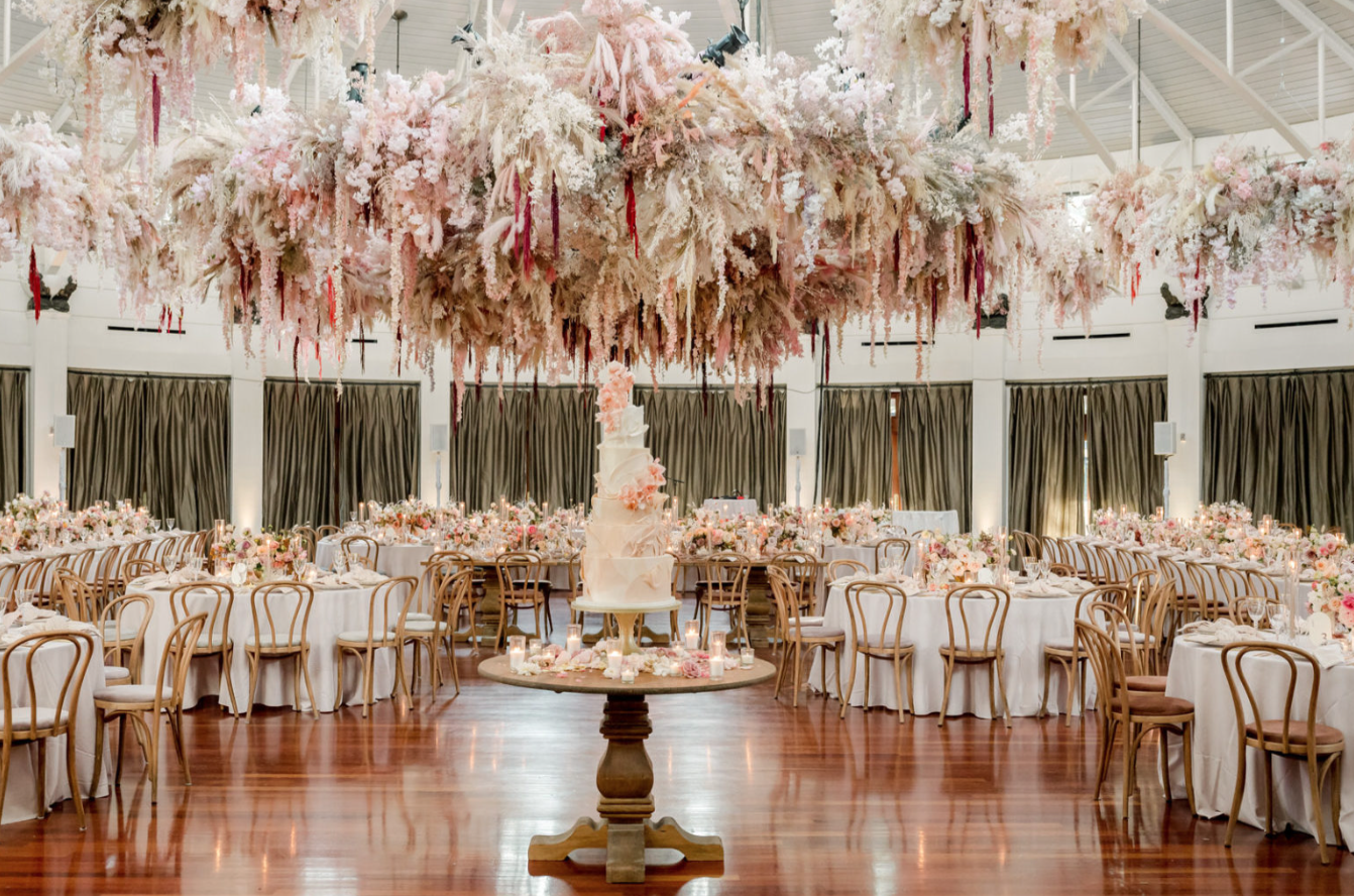 A wedding reception hall decorated with abundant pink and white floral arrangements, candles, and a multi-tiered wedding cake on a pedestal table, with round tables and wooden chairs arranged around the room.