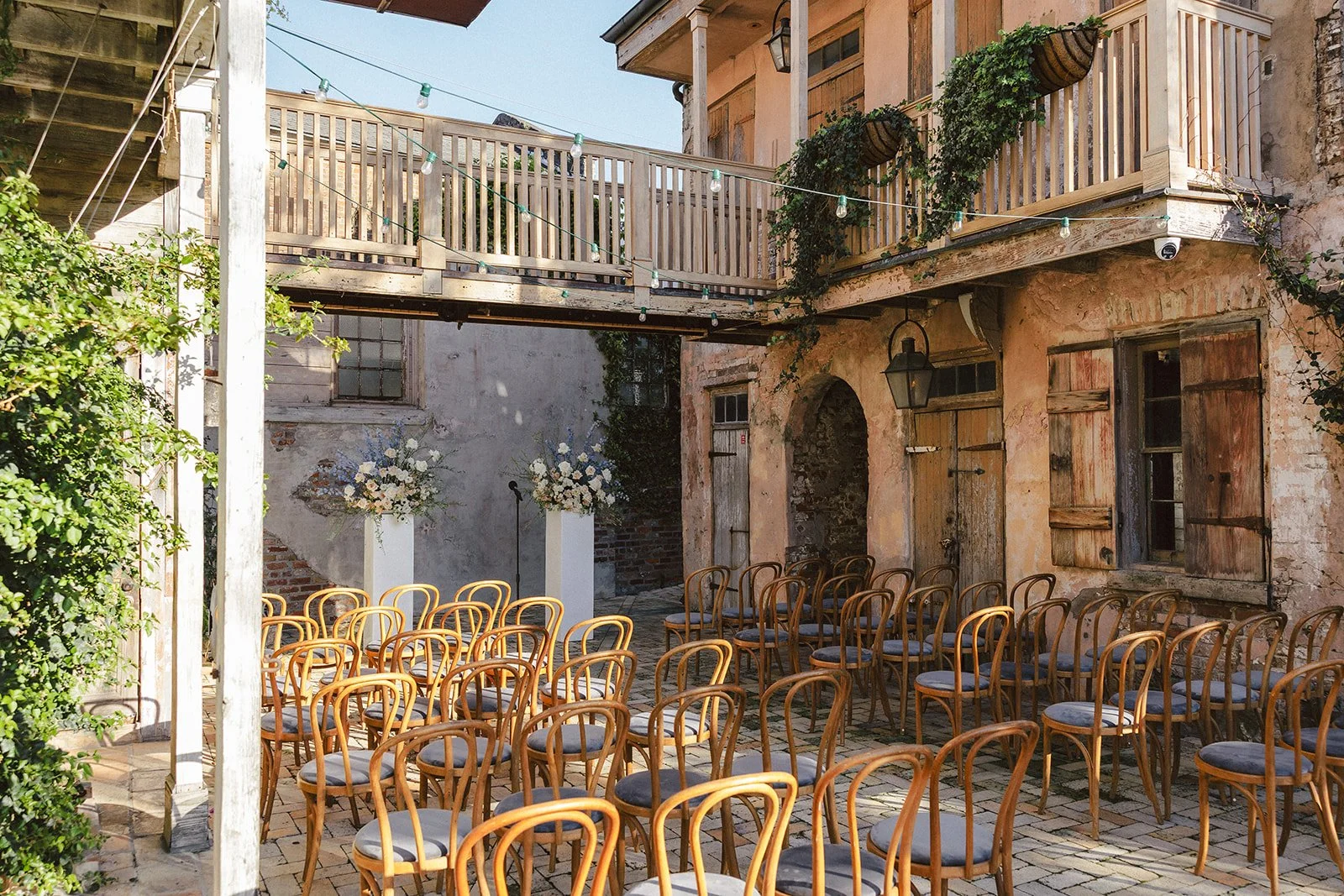 An outdoor courtyard set up for an event, with rows of wooden chairs with gray cushions facing a small stage with floral arrangements, surrounded by rustic brick and stucco buildings and a wooden balcony overhead.