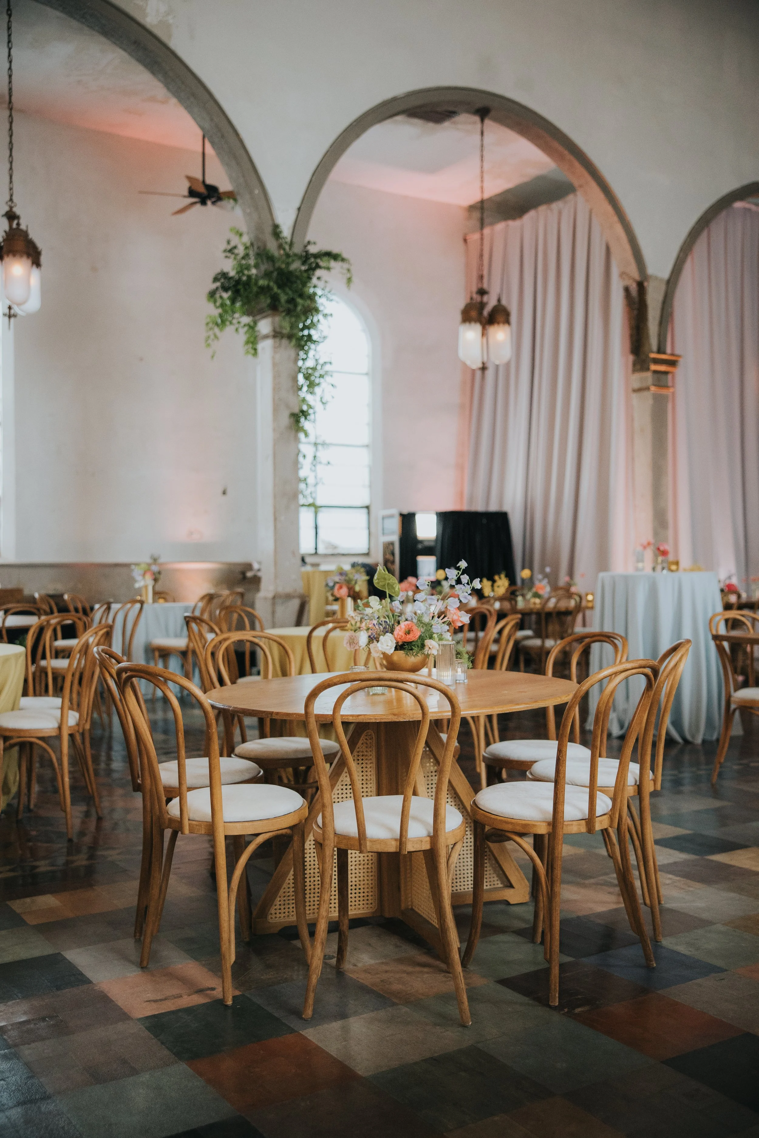 A decorated banquet hall with round tables covered in white and yellow tablecloths, each with floral centerpieces, wooden chairs, and a high, arched ceiling with hanging lights and curtains.
