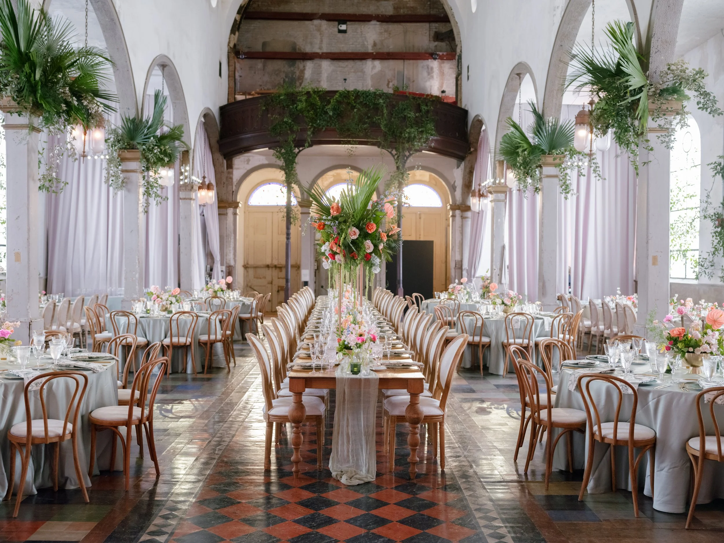 A large banquet hall decorated with floral arrangements, featuring a long dining table and classical architecture with a high arched ceiling, white curtains, and greenery accents.