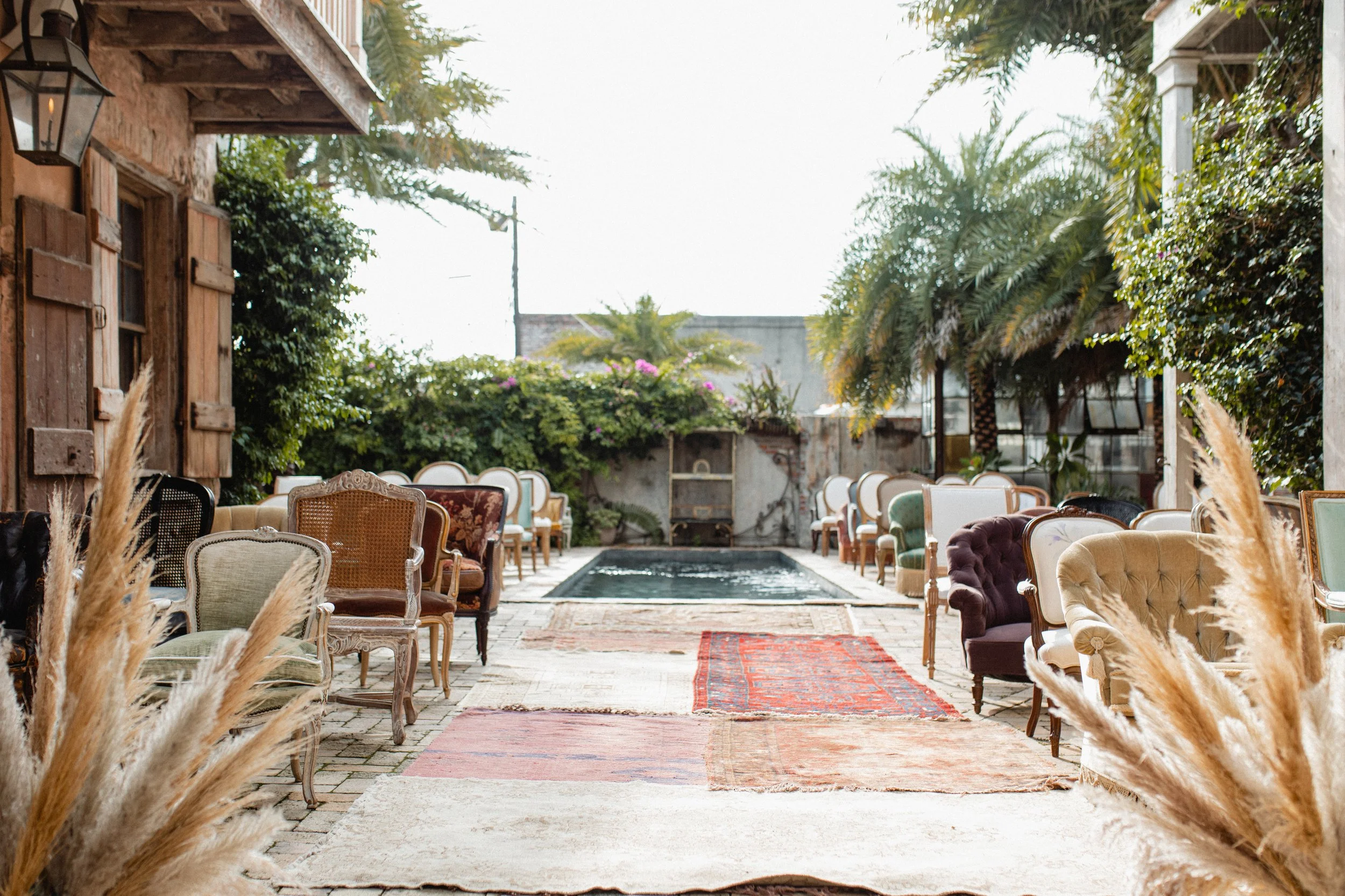 An outdoor seating area with vintage-style chairs and sofas arranged along a brick pathway, with a small pool in the center, surrounded by lush greenery and potted plants. The area is shaded by a wooden building and trees, with sunlight filtering thr