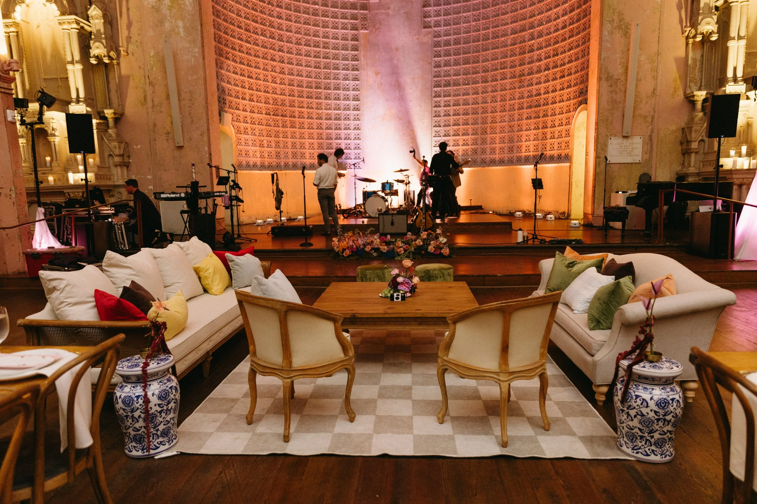 A stage set up for a musical performance inside a grand hall with ornate architecture, soft purple and warm yellow lighting. Musicians are preparing, with musical equipment and microphones on stage, fronted by a floral arrangement. Seating with color