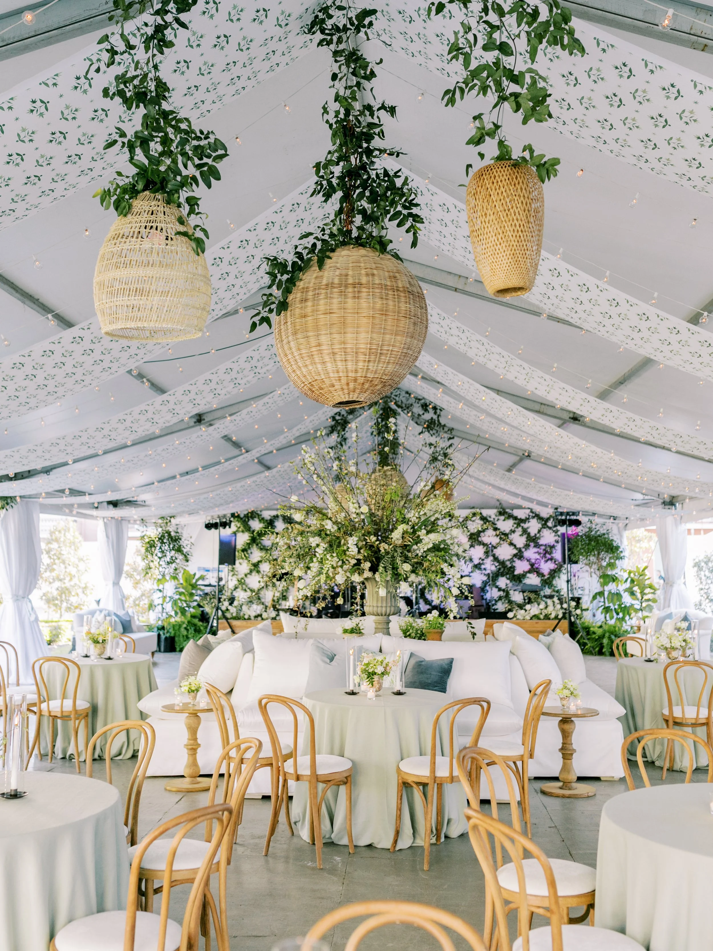 Decorated indoor event space with white chairs and round tables, large floral centerpiece, hanging wicker lanterns, and ceiling draped with white fabric and green leaf prints, string lights overhead.