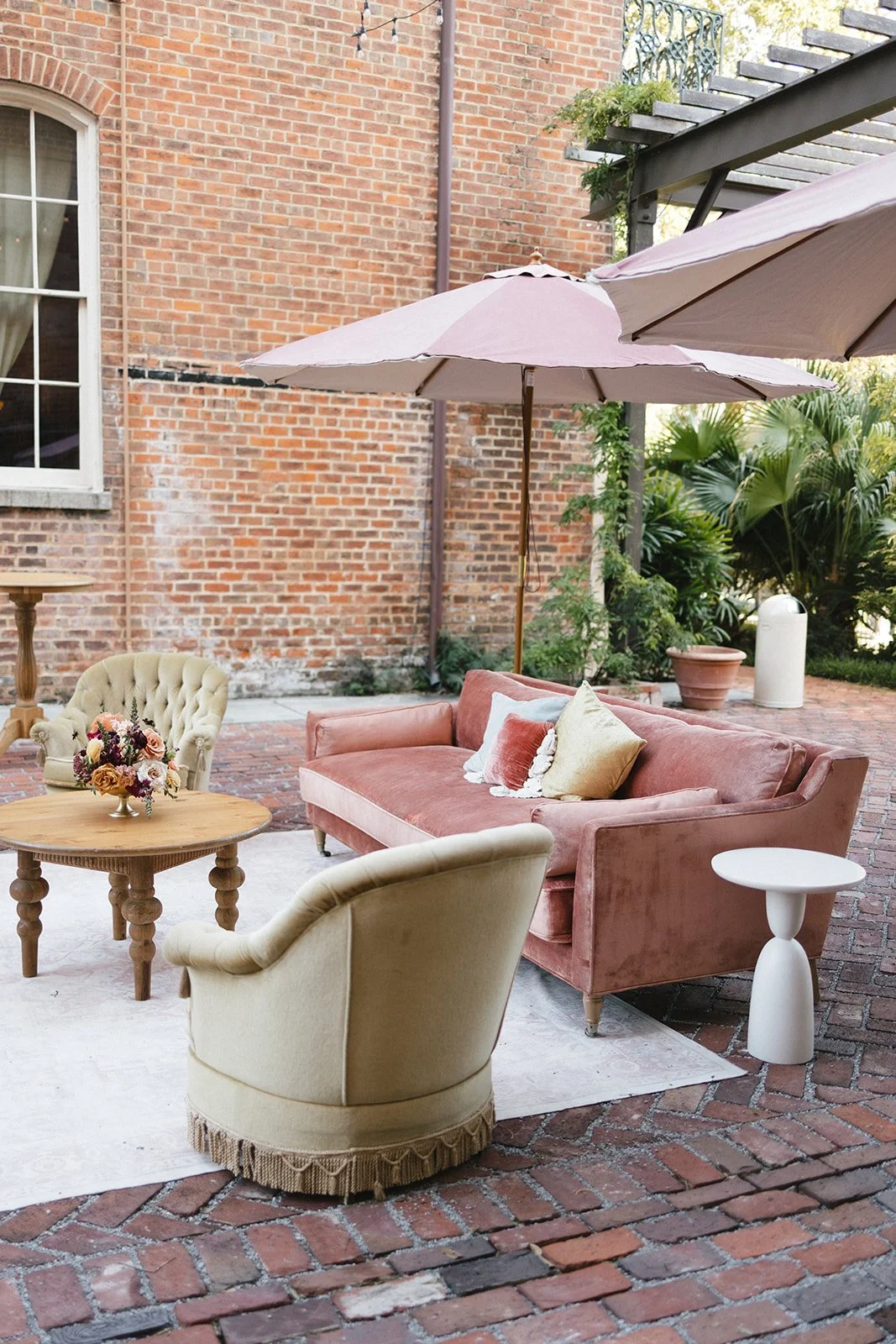 Outdoor seating area with pink velvet sofa, beige armchair, small round wooden table with flower arrangement, pink and beige umbrellas, brick patio, potted plants, and a brick building wall in the background.