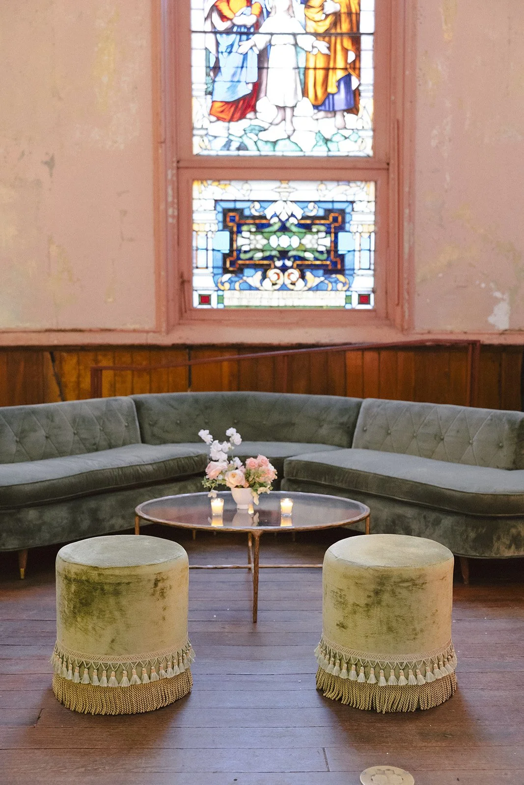 Vintage chapel interior with a curved green velvet sofa, two pale yellow velvet ottomans with fringe trim, a round wooden coffee table with a flower arrangement and candles, a stained glass window depicting religious figures, and wooden wall paneling