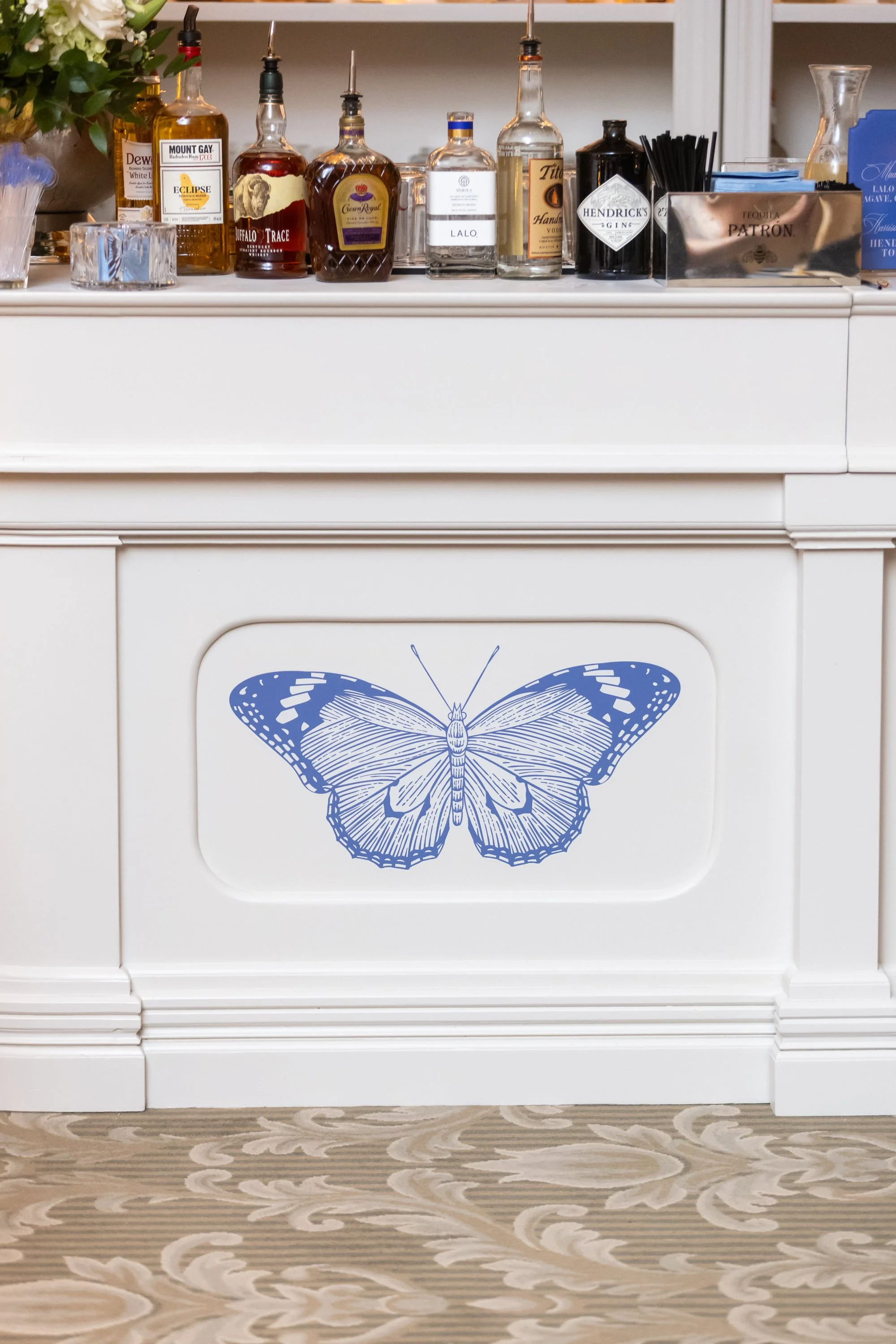 A white bar with a large blue butterfly graphic on the front panel, topped with bottles of alcohol and bar accessories, and a floral arrangement to the left.