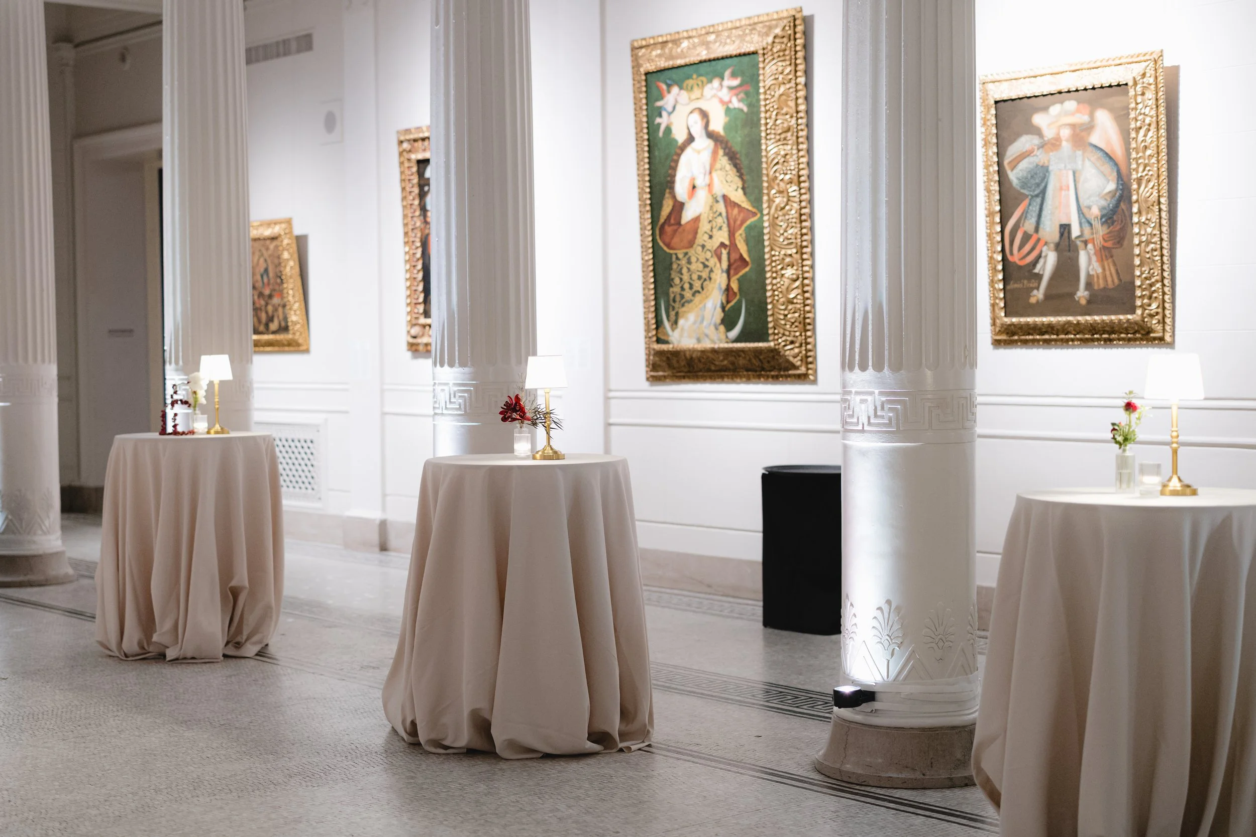 Art gallery with framed paintings, round tables with white tablecloths, small lamps, and decorative flowers, in a bright, elegant space with white columns.