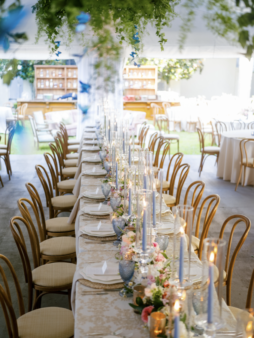 A long banquet table decorated with pink and white flowers, blue candles, and elegant glassware, set up outdoors under a tent with green leaves and hanging decorations.