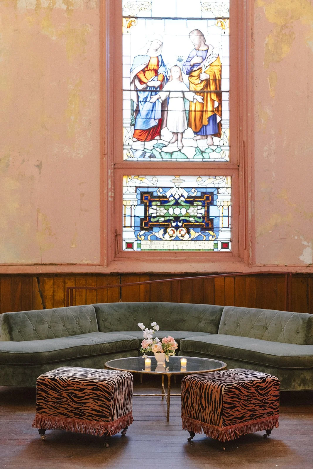 A stained glass window depicts a religious scene with three figures, including a child, in a church interior. Below the window, a vintage green velvet sofa and two zebra-print ottomans surround a glass coffee table with a flower arrangement and candl