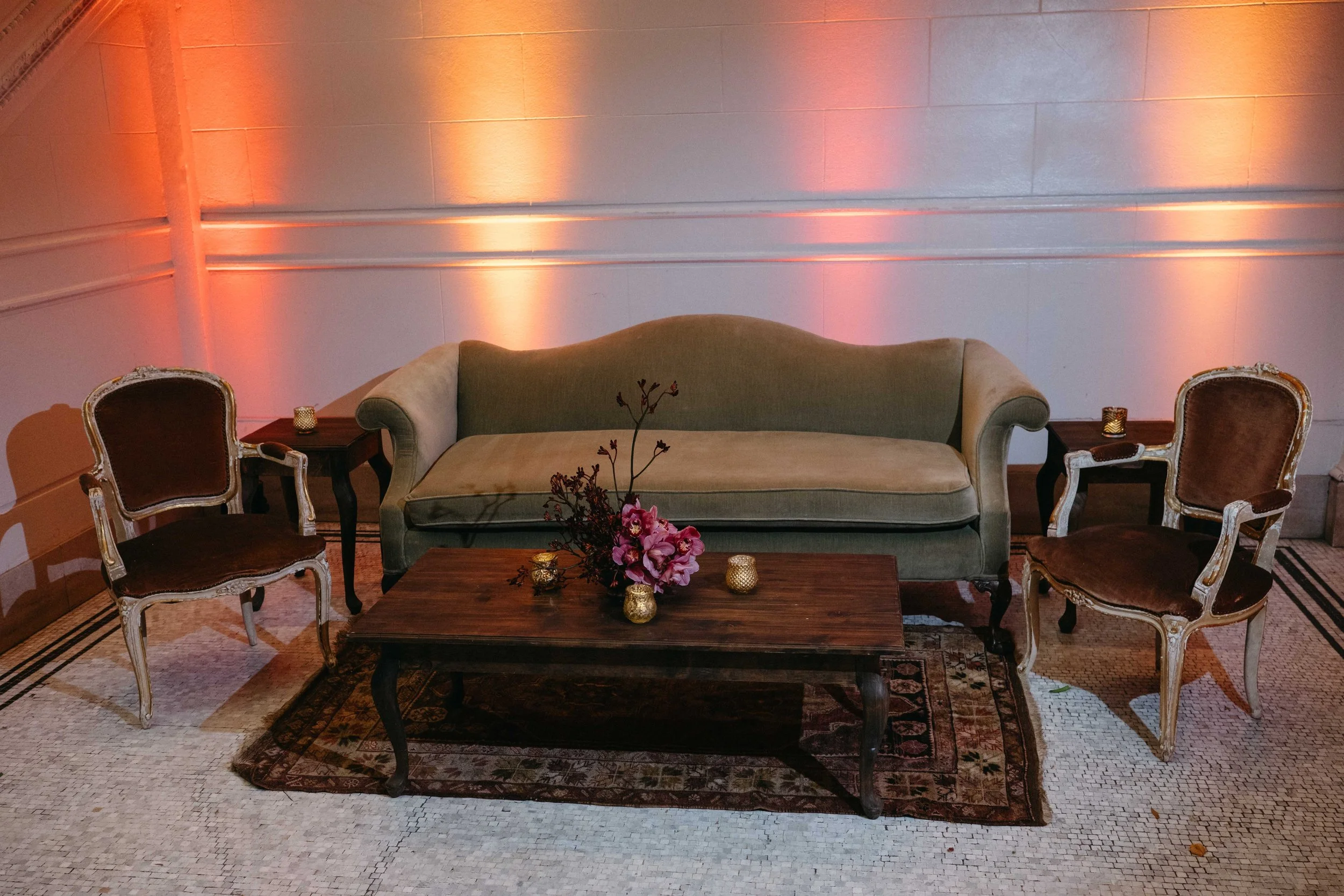 A vintage-style living room with a beige sofa, two antique chairs with carved wooden frames, a wooden coffee table with pink and purple flowers and small candle holders, against a wall illuminated with orange and pink ambient lighting, with a pattern