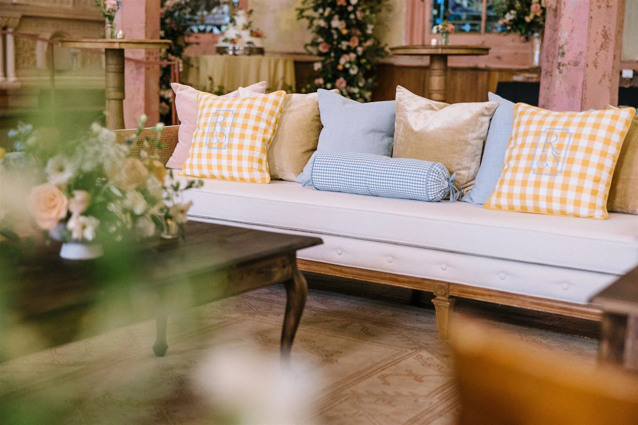 A cozy indoor lounge area with a white sofa adorned with an assortment of pillows in pastel colors and checkered patterns. There is a wooden table with a floral arrangement in the foreground. The background features wooden paneling, pink painted bric