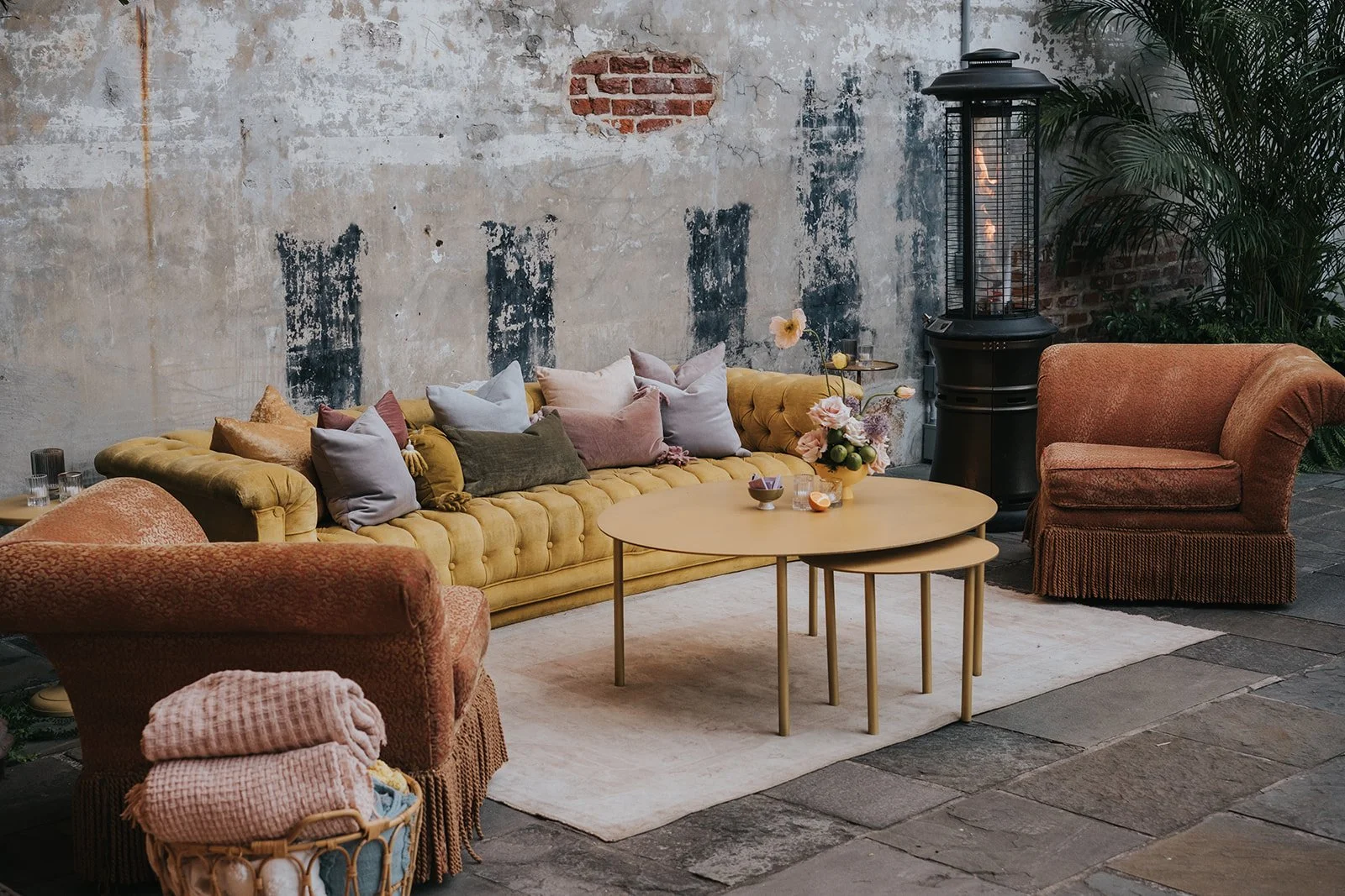 A cozy living room with a yellow velvet tufted sofa, an orange armchair, and a peach-toned armchair on a light rug. There are decorative pillows on the sofa, a small stack of folded blankets in a basket, and a round coffee table with small decorative