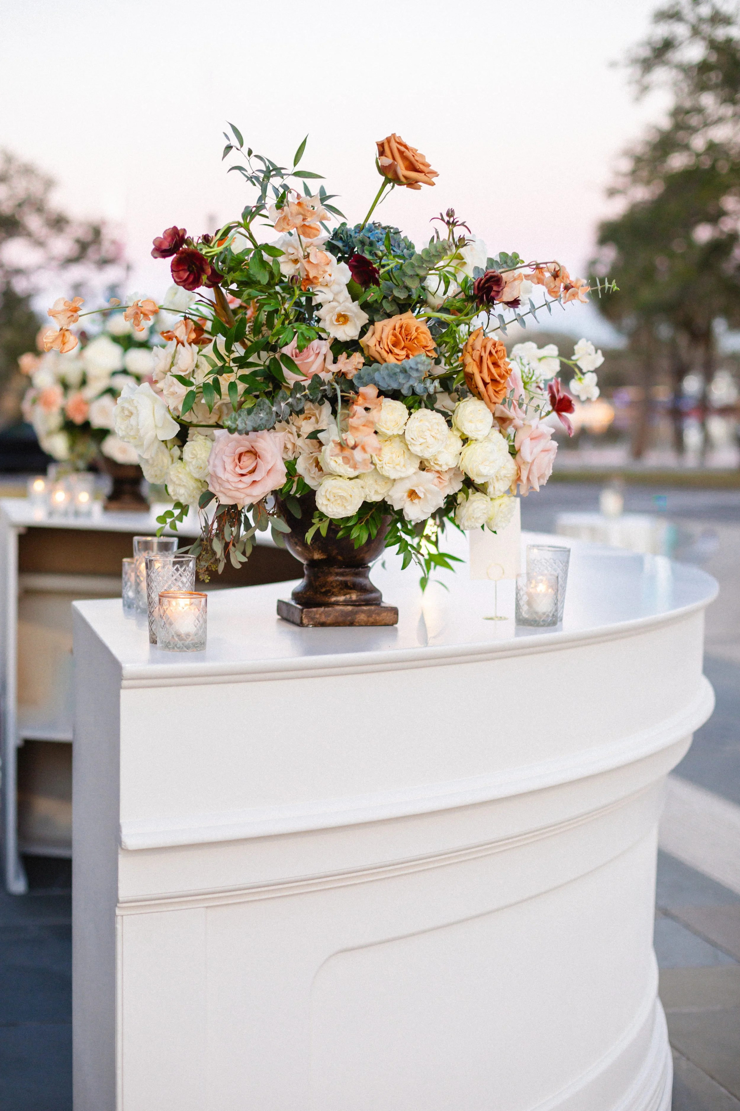 A floral arrangement on a white curved table with candles in glass holders, outdoors with blurred background trees and sky at sunset.