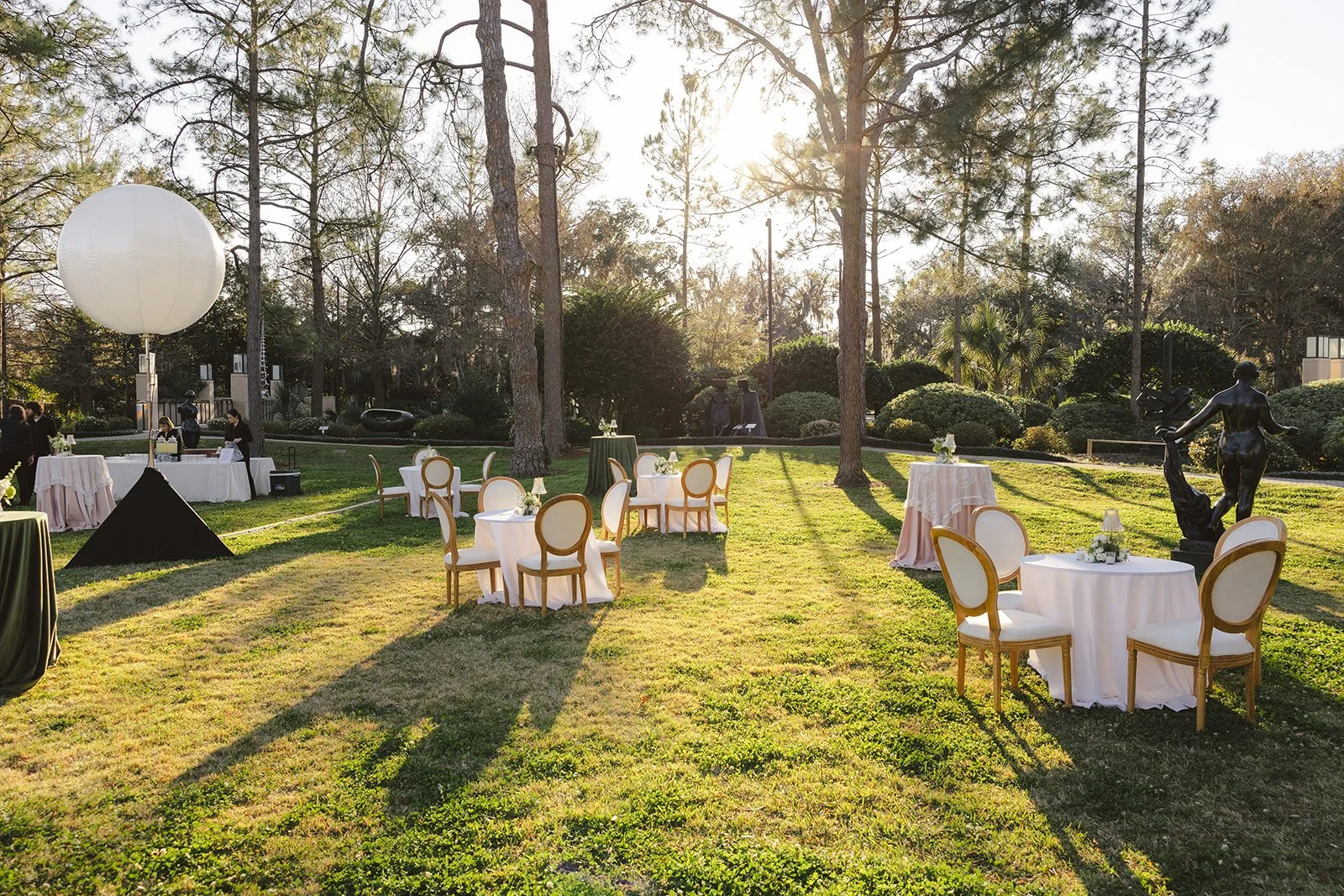 Outdoor event setup with round tables covered in white tablecloths and gold-trimmed chairs, statues, a large white balloon, and lush greenery in a park-like setting during sunset.
