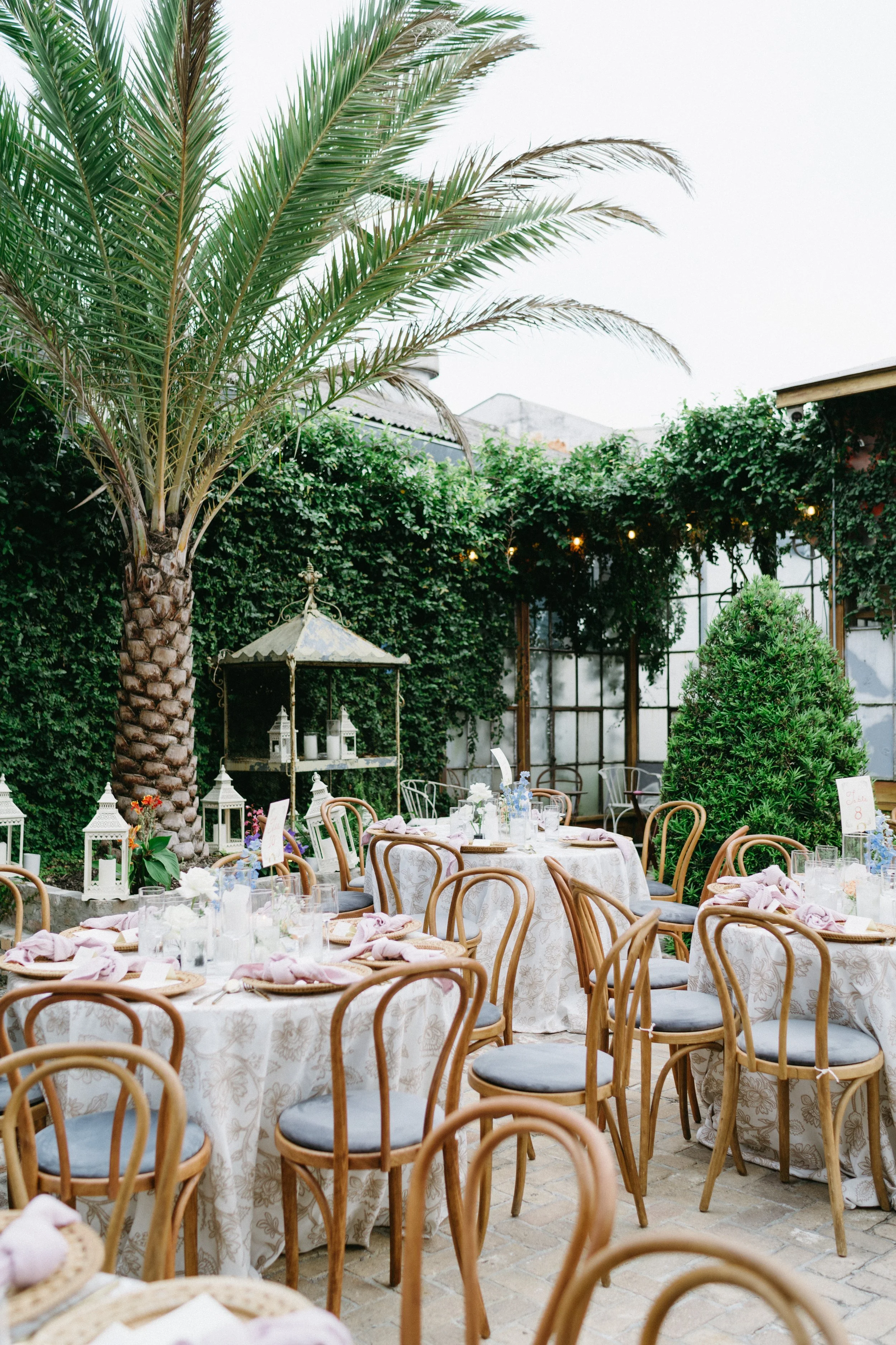 Outdoor garden decorated for a celebration with round tables covered in white floral tablecloths, surrounded by wooden chairs with gray cushions, adorned with pink napkins and floral centerpieces, set amidst lush greenery, including a large palm tree