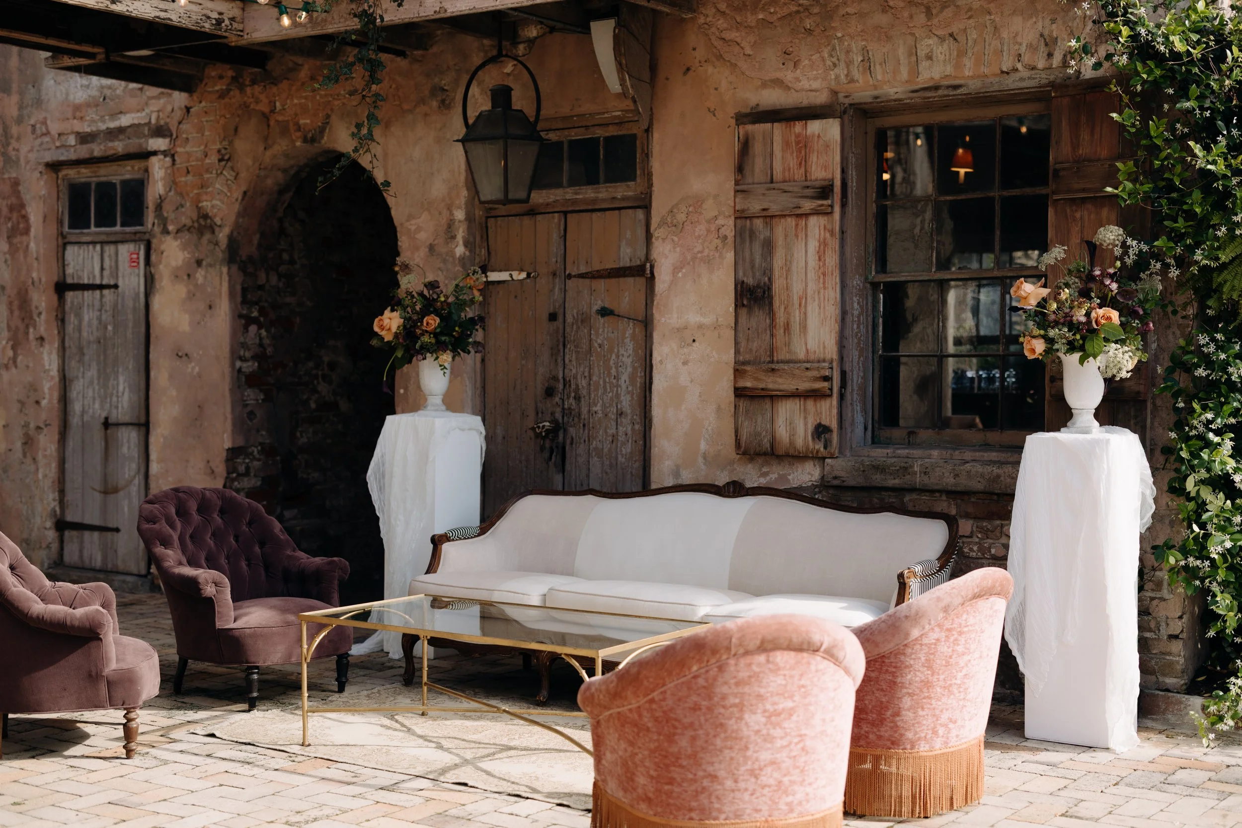 A cozy indoor space with vintage furniture including velvet armchairs and a white sofa, decorated with floral arrangements in tall vases, set against a rustic wall with wooden shutters, a window, and exposed brick.