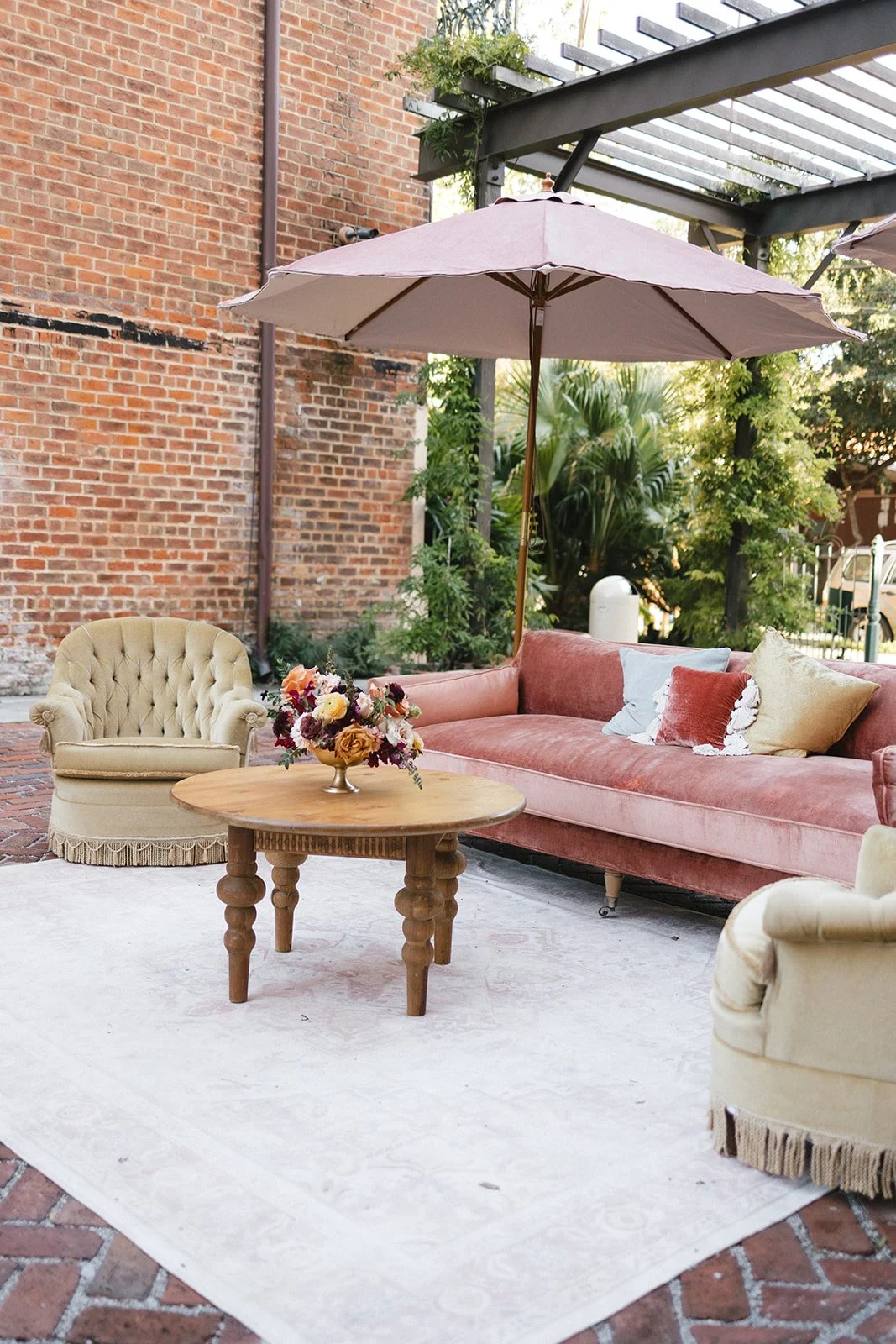 Outdoor patio with pink velvet sofa, cream tufted armchairs, wooden coffee table with a flower arrangement, large pink umbrella, and greenery, against a brick wall.
