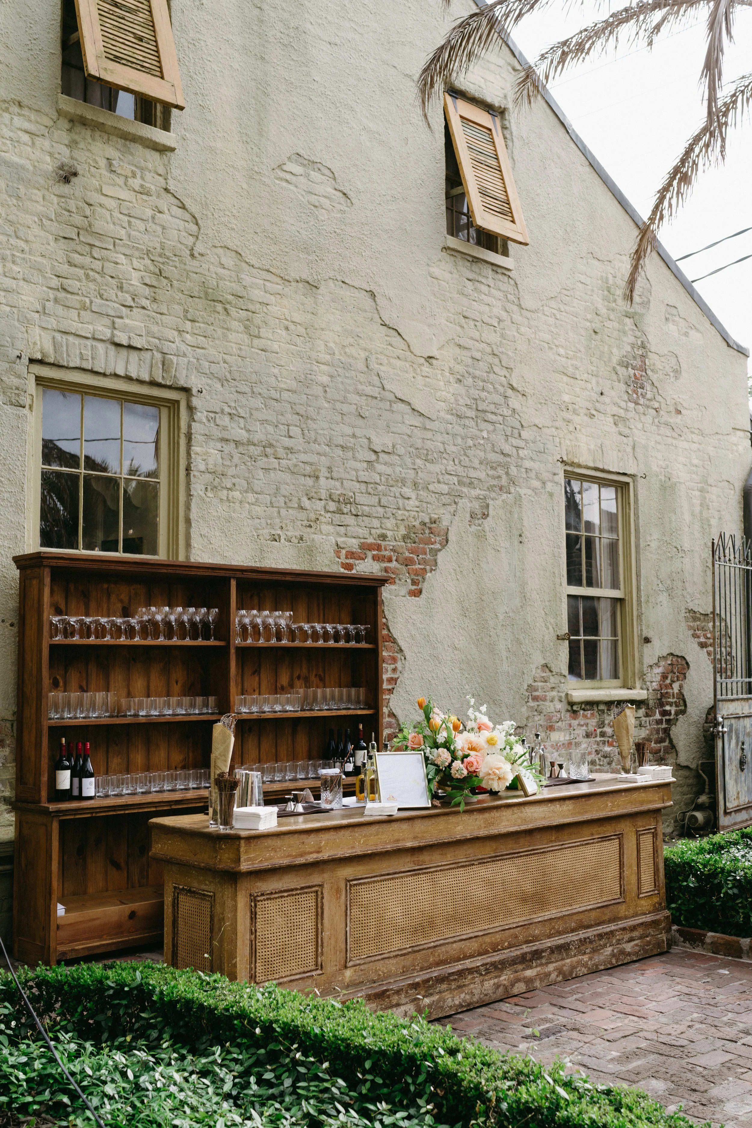 An outdoor rustic bar setup in front of an old brick and stucco wall with peeling paint, surrounded by greenery and a brick pathway. The bar surface holds a large floral arrangement, wine bottles, glasses, and bar accessories. A wooden shelving unit 