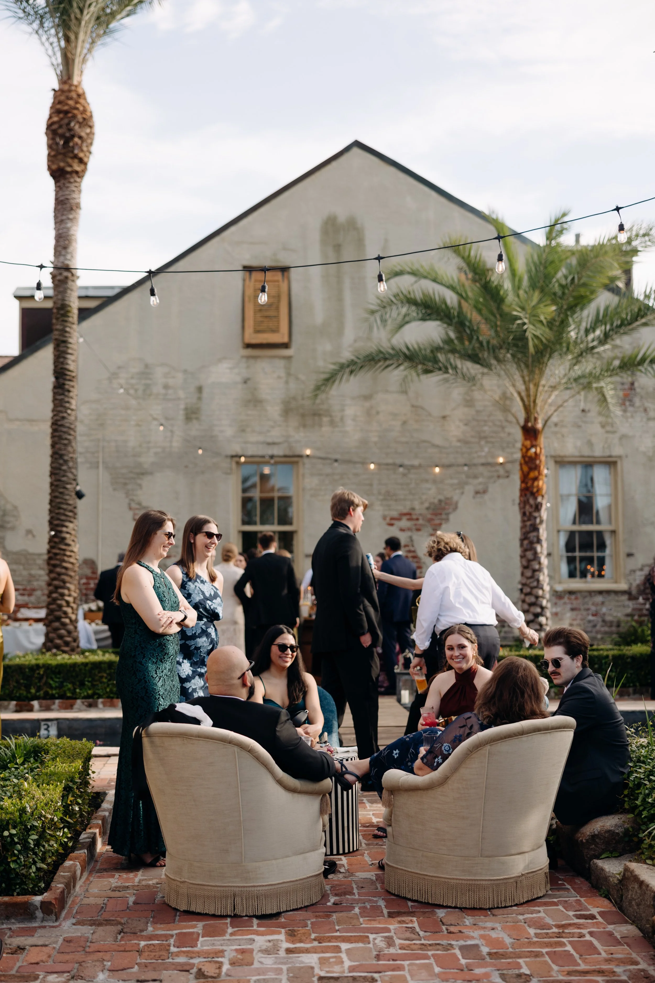 People socializing outdoors at a gathering, with palm trees and string lights in the background, sitting in chairs and standing in conversation.
