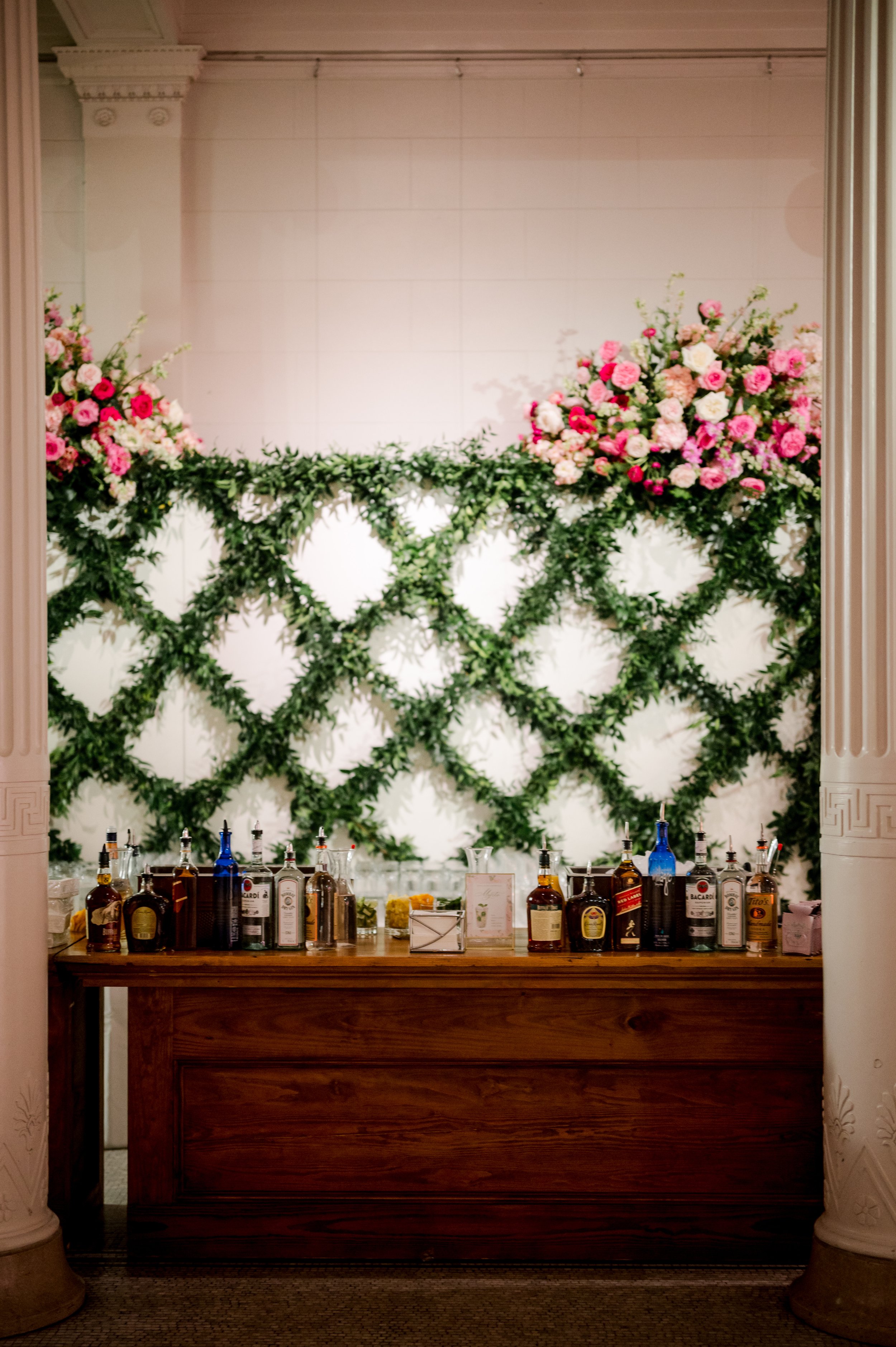 A bar with various bottles of alcohol on a wooden counter, behind which is a large floral arrangement with pink and white flowers and green foliage mounted on a white wall.