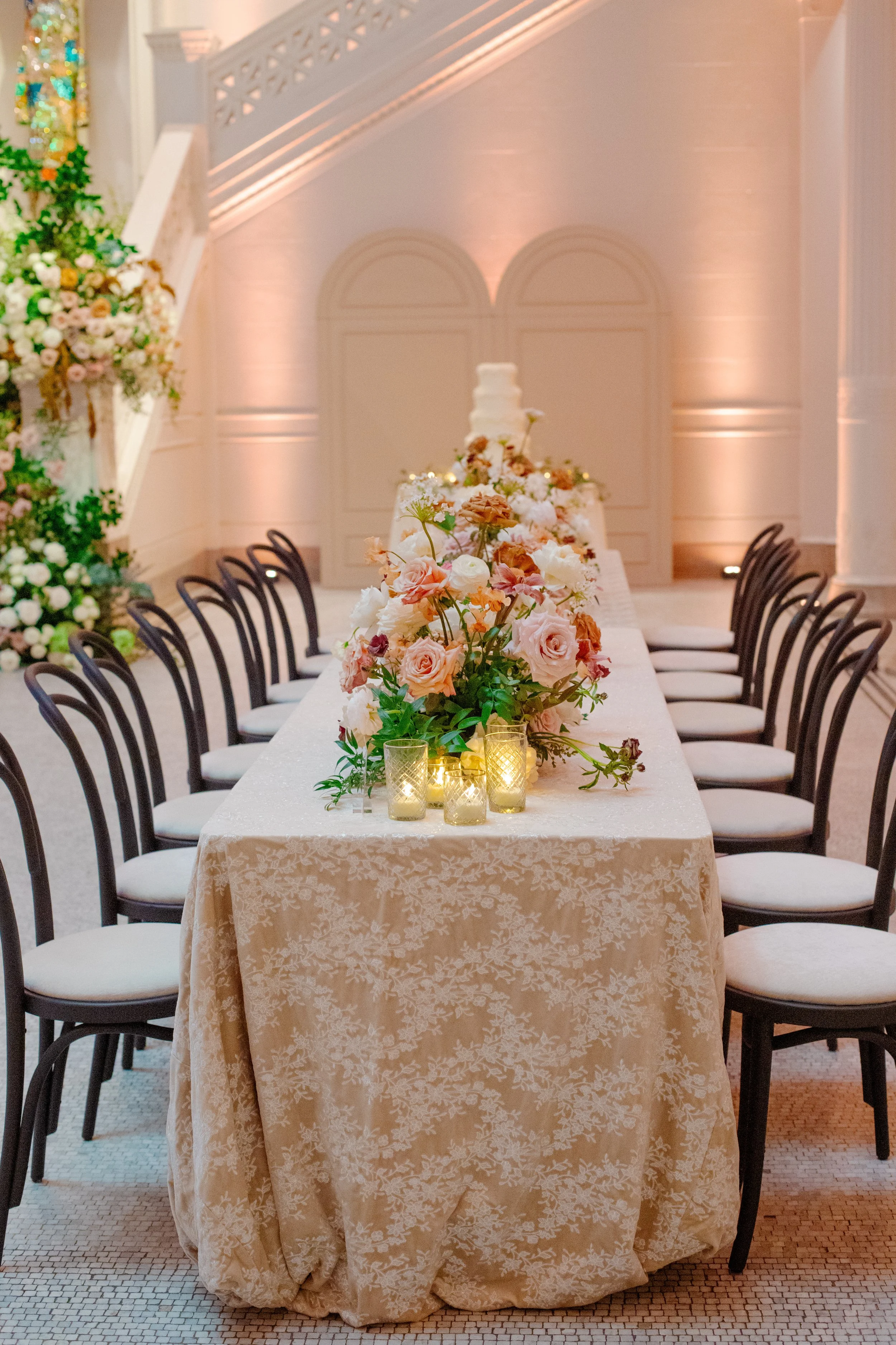 A long rectangular table with a cream-colored lace tablecloth, floral centerpiece with pink and white roses, and small candles in glass holders. The table is surrounded by black chairs with white cushions, and in the background, there is a wall with 