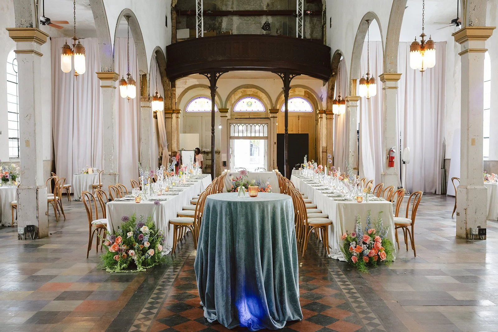 Elegant banquet hall with long white-draped tables, floral centerpieces, surrounded by wooden chairs, and high arched windows draped with white curtains, in a historic building with exposed stone walls and ceiling fans.