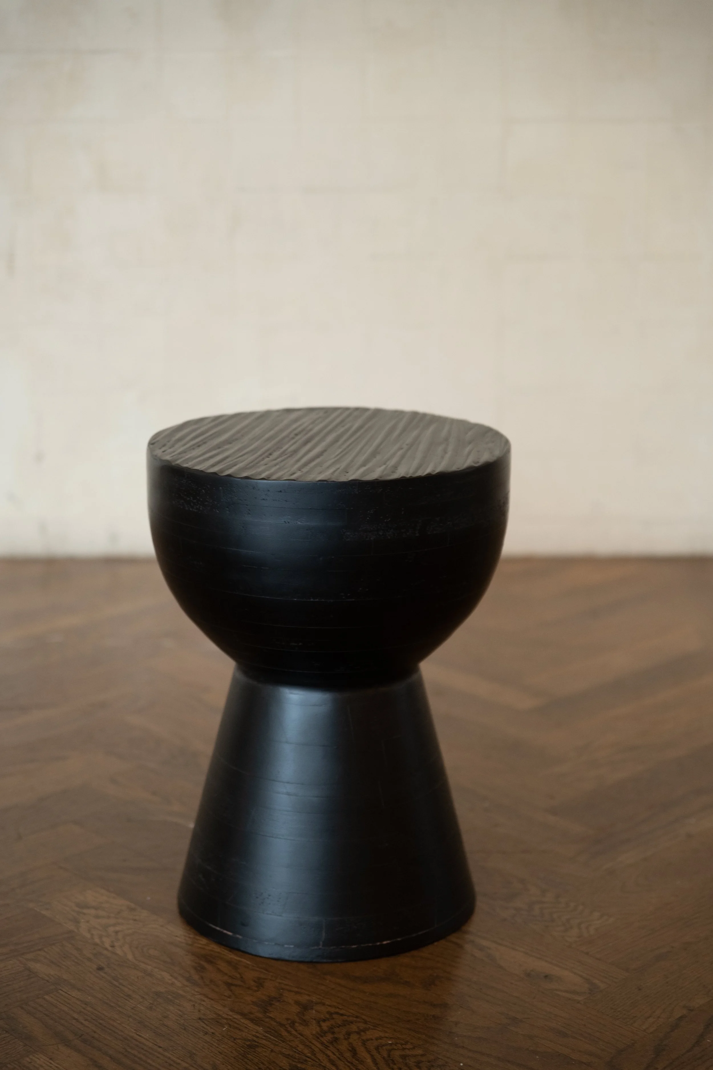 A black wooden stool with a round top and a conical base positioned on a wooden floor, against a plain light-colored wall.