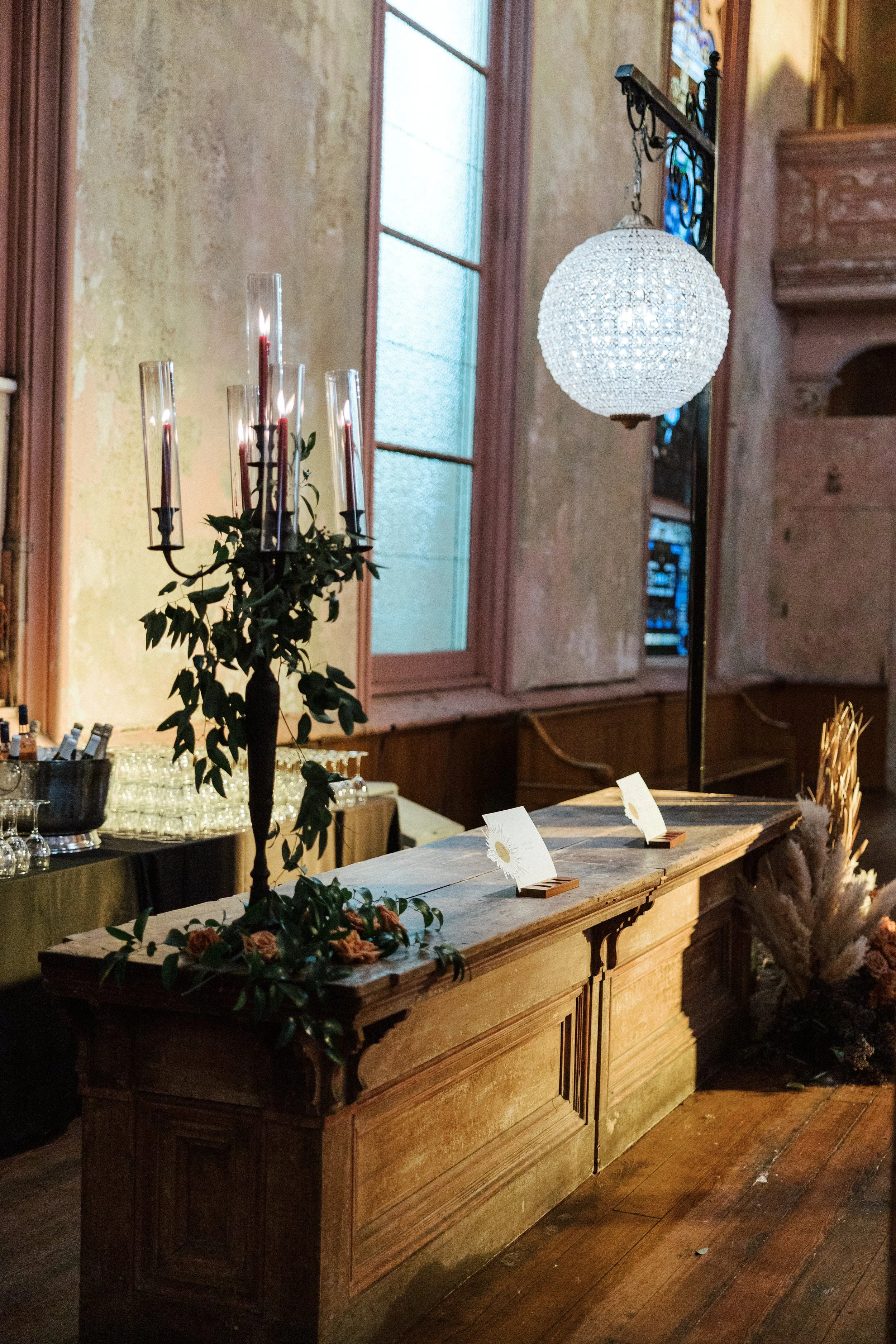 Vintage wooden counter with white tablecloth, floral decoration, and small signs, inside a rustic building with large window, stained glass, and hanging chandelier.