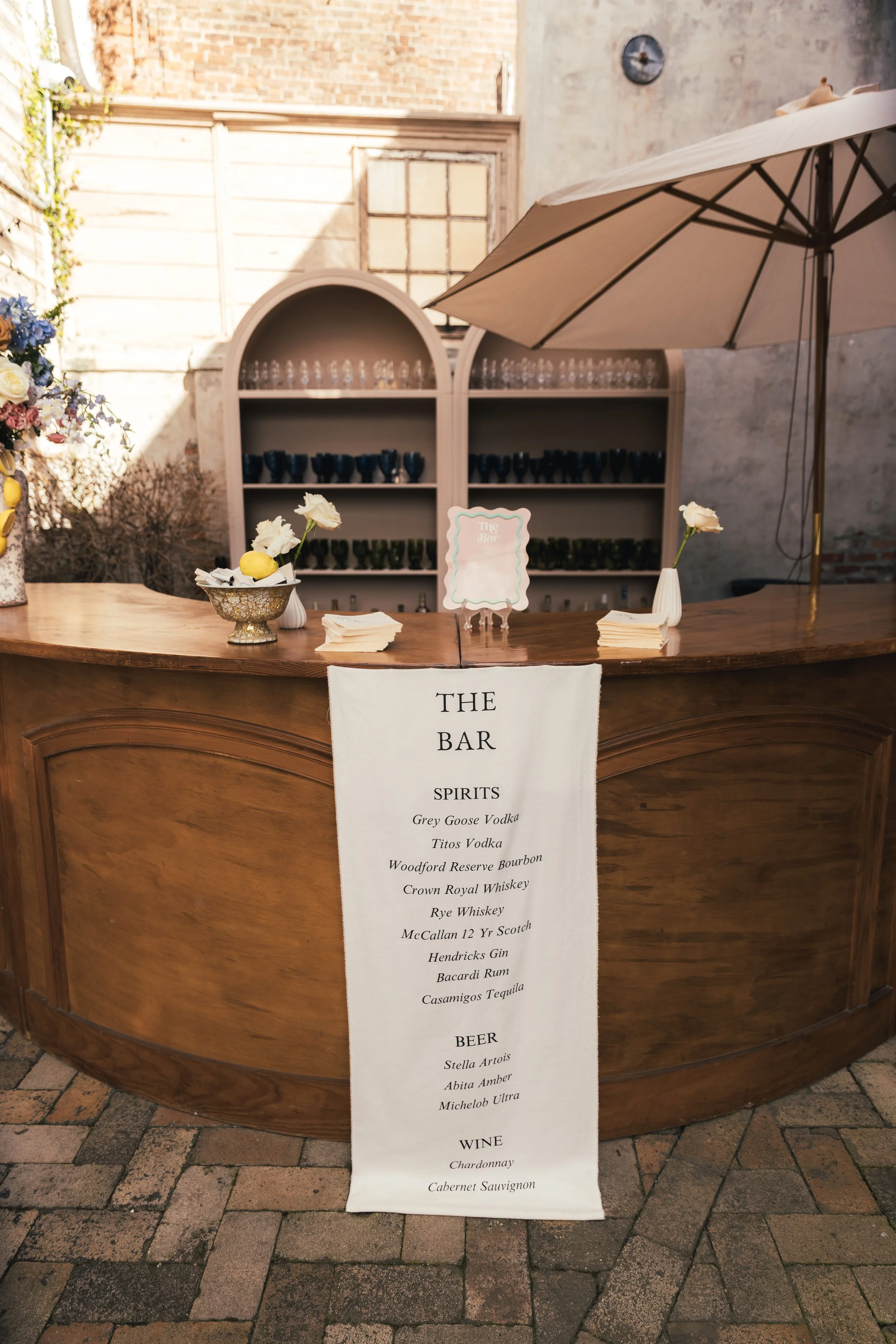 A wooden bar counter with a menu list hanging from it, listing spirits, beer, and wine options. There are white flowers in vases, a bowl with lemons and flowers, and a large patio umbrella providing shade. In the background, there are shelves with gl