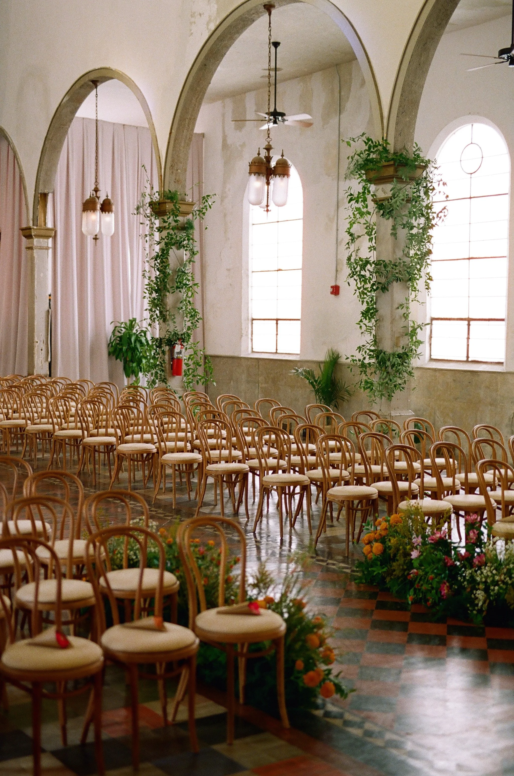 Indoor space with rows of light wooden chairs with white cushions, decorated with flowers, large arched windows, hanging vintage-style lamps, green plants in planters, and warm natural lighting.