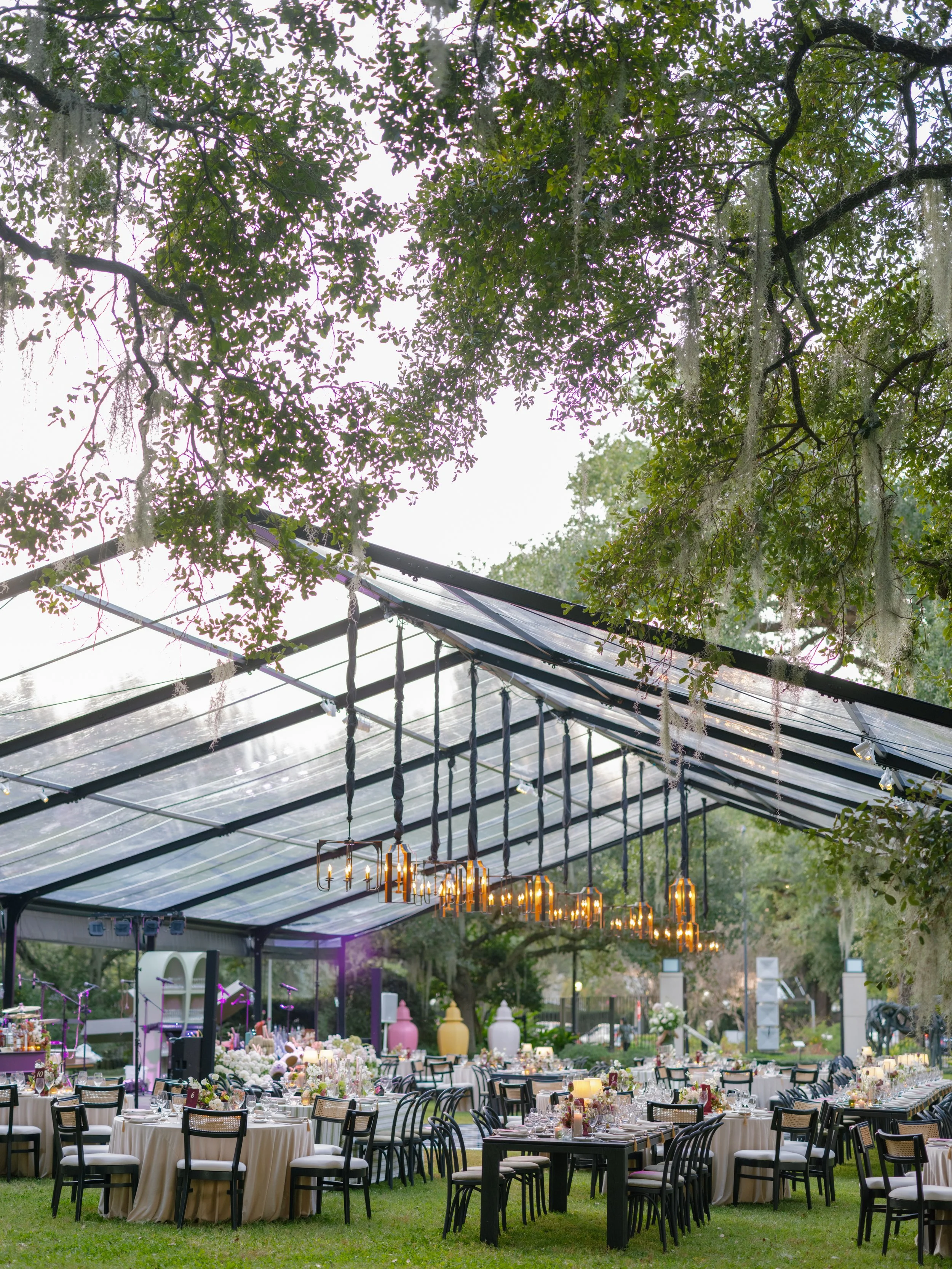 An outdoor wedding reception under a clear tent with decorated tables, chandeliers hanging from the ceiling, and trees in the background.