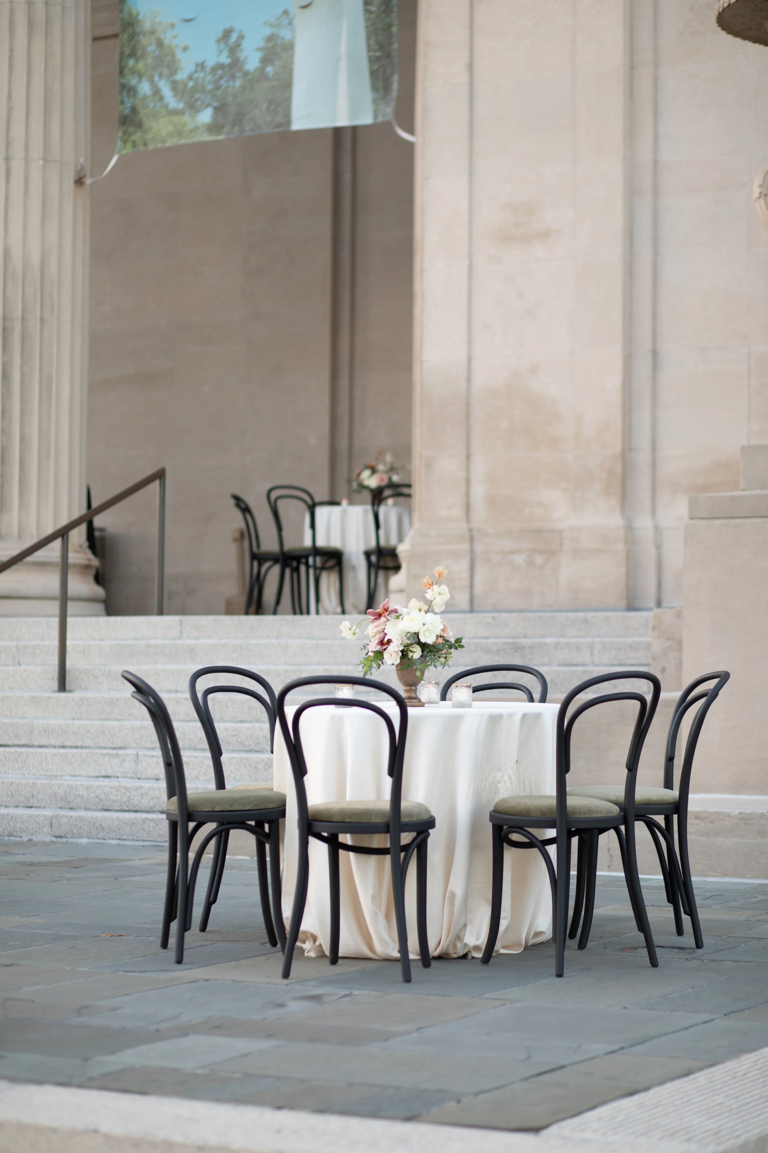 Elegant outdoor dining setup with a round table, white tablecloth, floral centerpiece, and black chairs, situated on stone steps in front of a historic building with large columns.