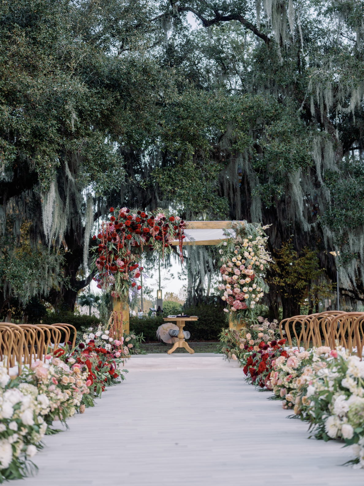 Outdoor wedding ceremony setup with floral decorations, a wooden arch, and chairs on a white walkway surrounded by trees.