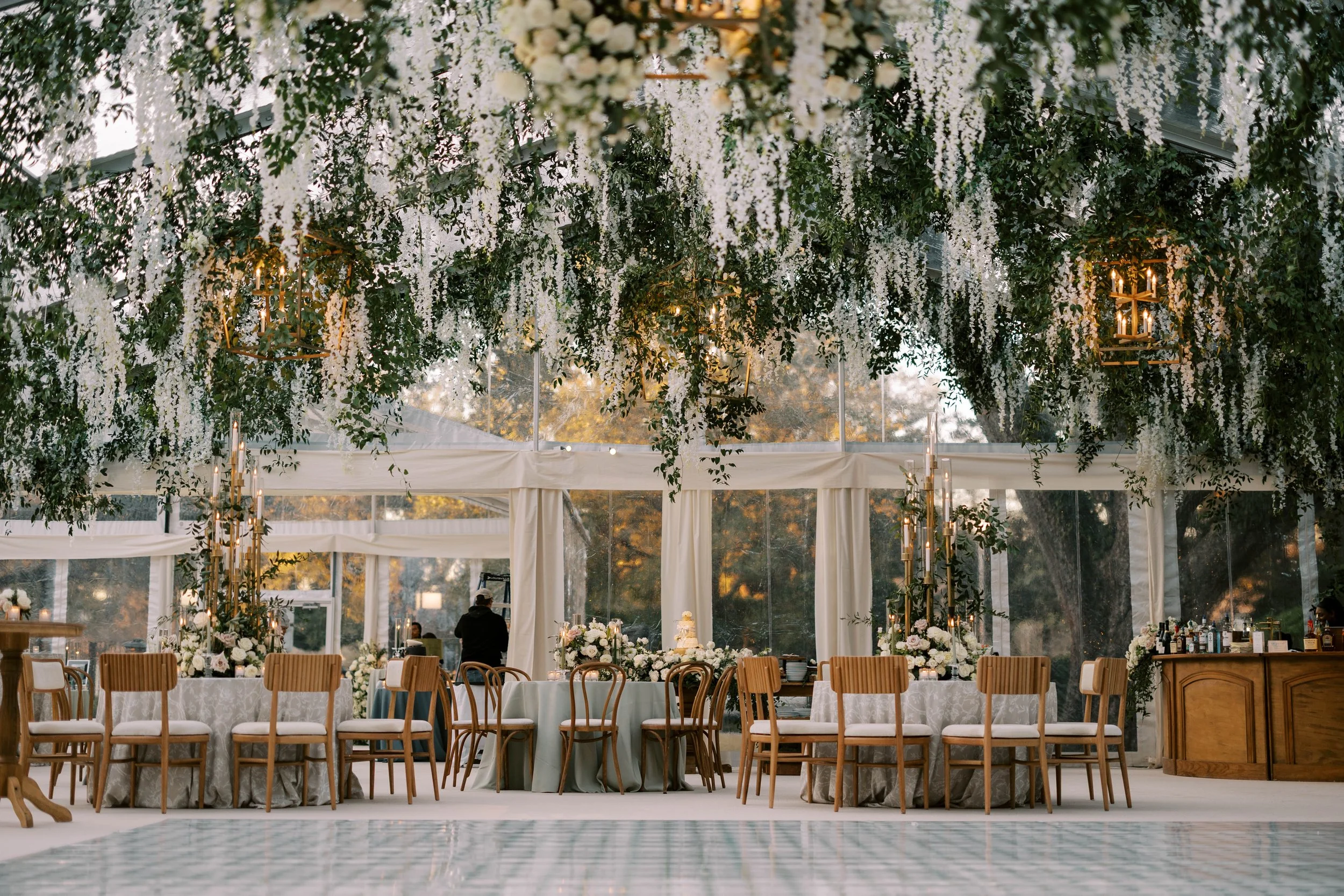 Elegant wedding reception setup with round tables, floral centerpieces, wooden chairs, and a decorated ceiling with hanging white flowers and greenery in a glass-enclosed venue.