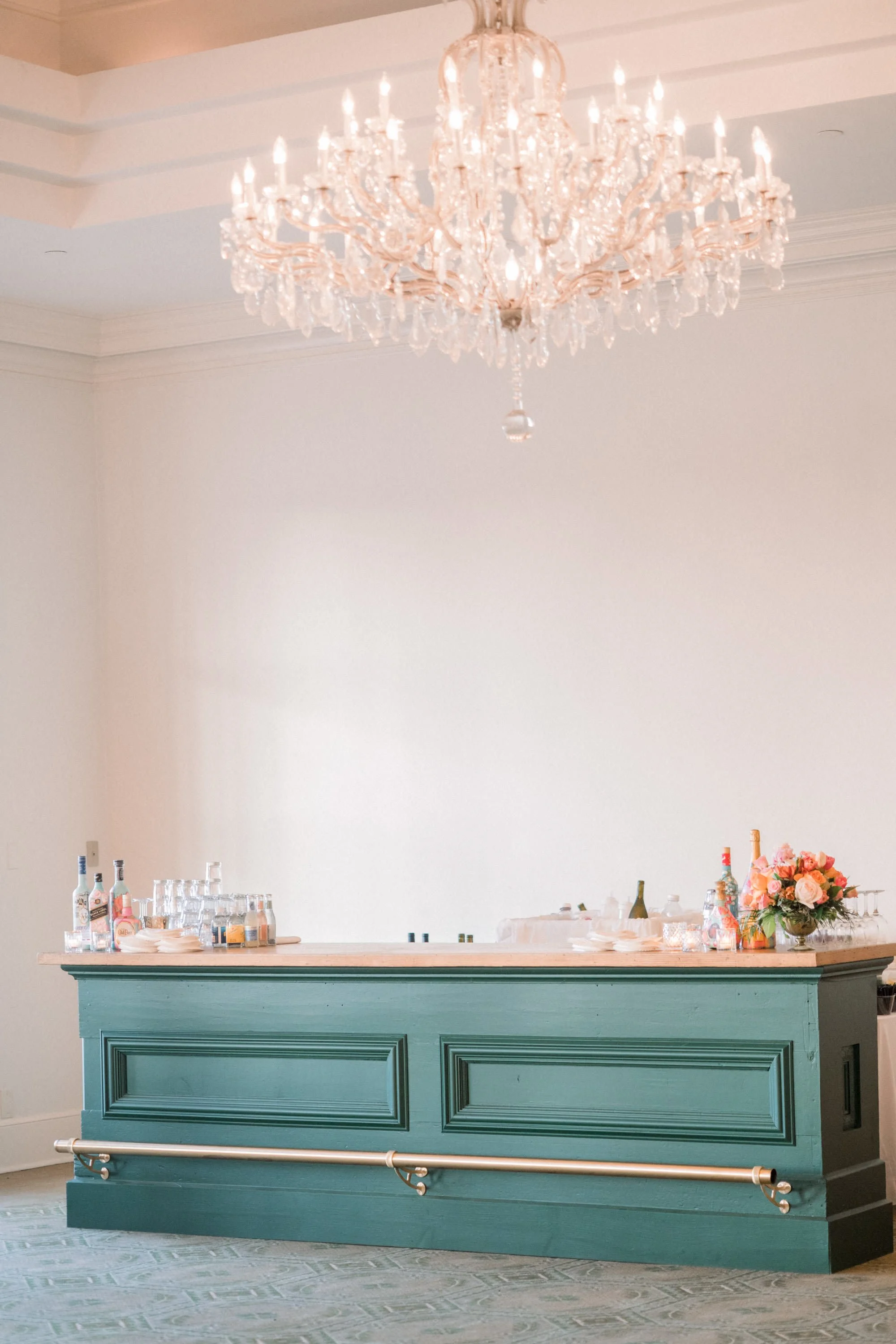 A teal-colored bar or counter with a wooden top, decorated with bottles, glasses, and a flower arrangement, situated beneath a large crystal chandelier.