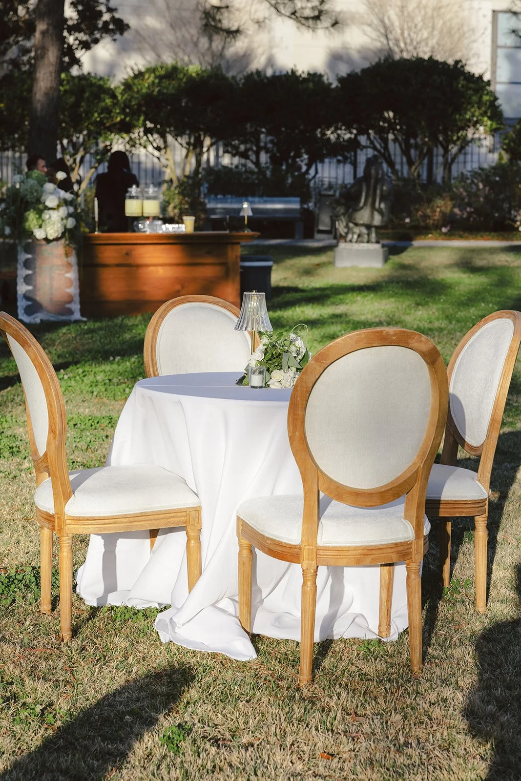 Outdoor event setup with a round table covered in a white tablecloth, four wooden chairs with white cushions, a small floral centerpiece, and a small table lamp, with a garden, statue, and people in the background.