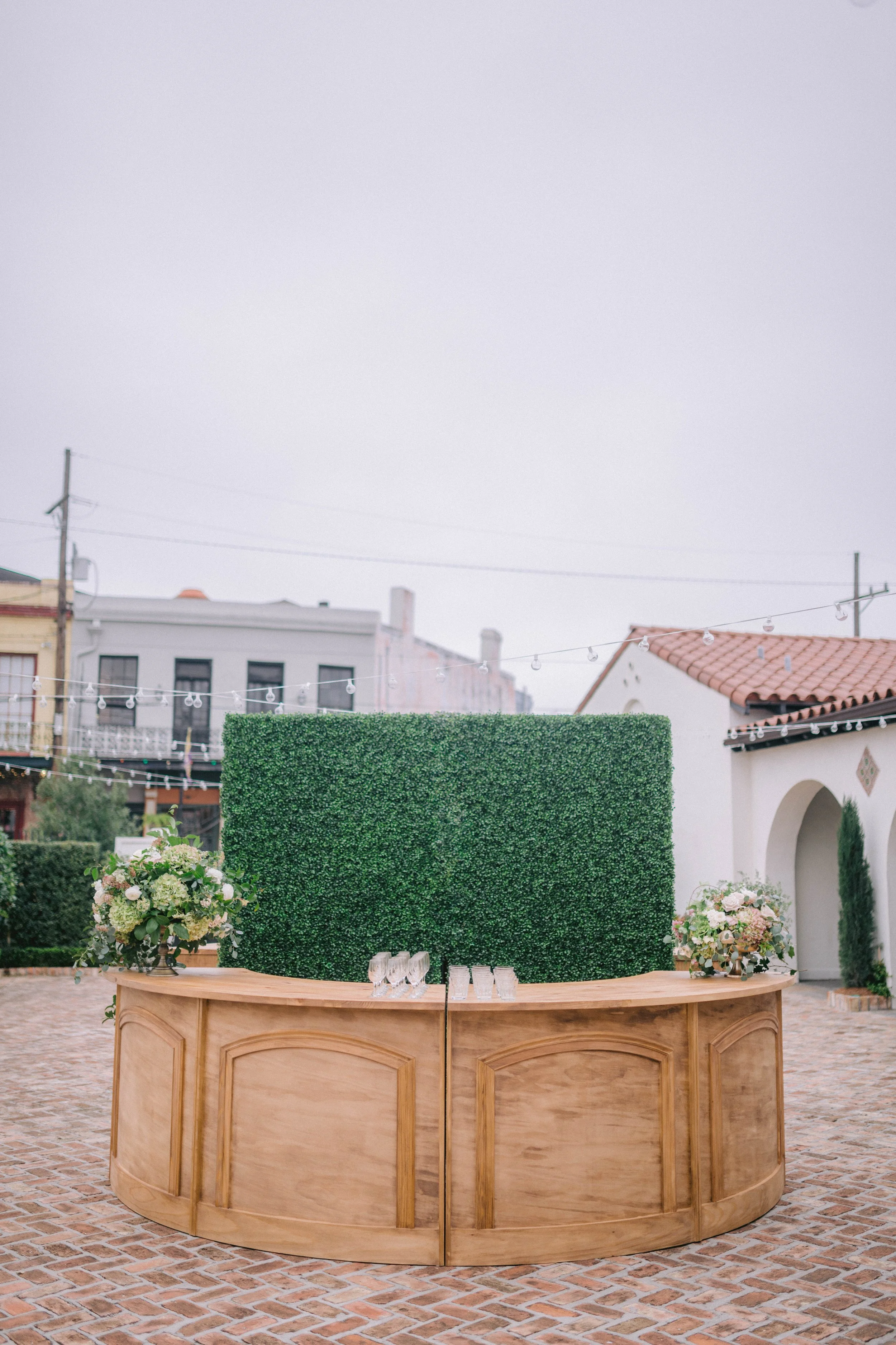 Outdoor event setup with a curved wooden bar, floral arrangements, a green hedge backdrop, and string lights, with buildings and a cloudy sky in the background.